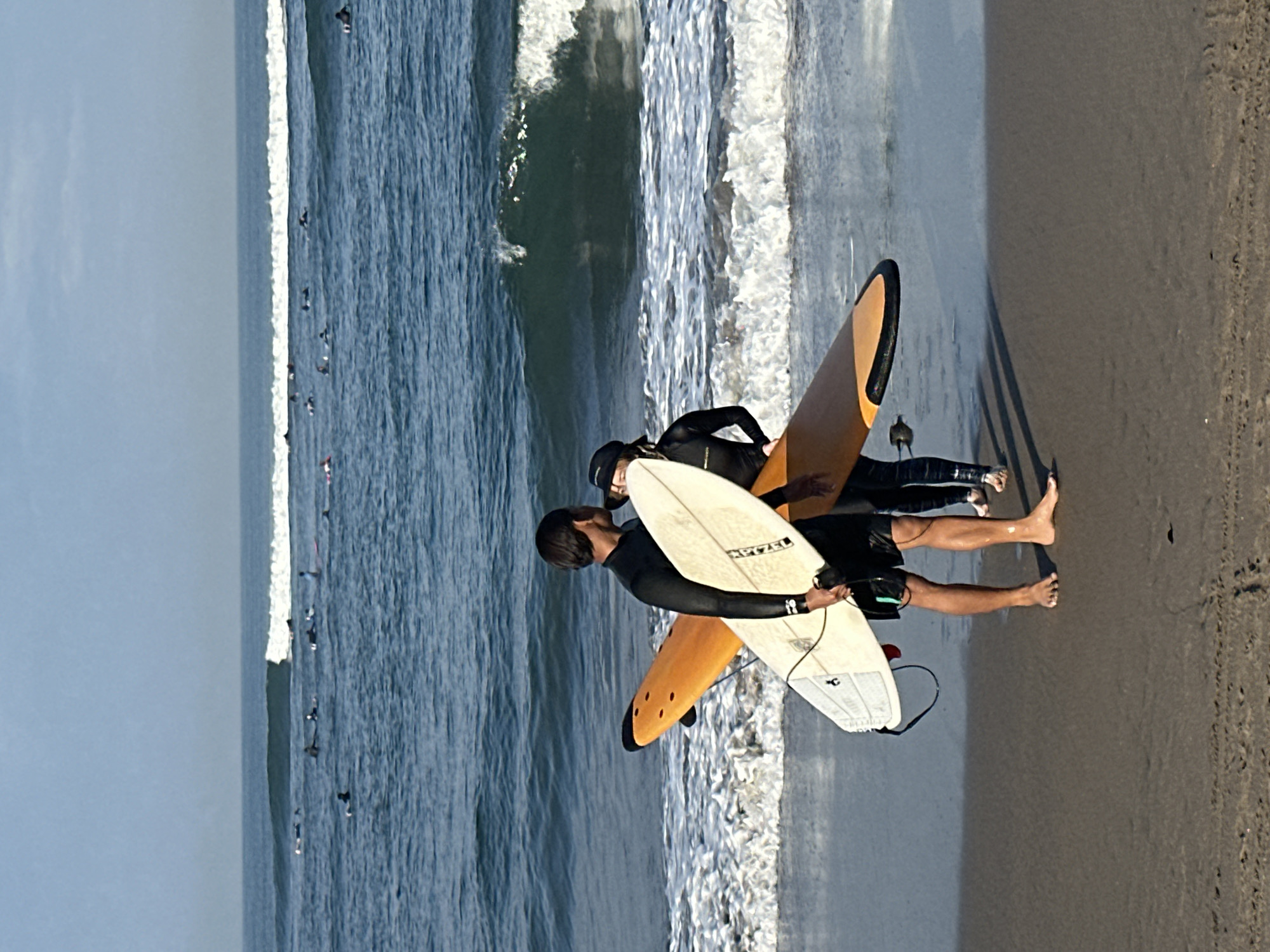 At the shore of North Kuta, Indonesia, two surfers stand on the wet sand, facing the ocean. The surfer in the foreground, a man with dark hair, wears a black wetsuit with shorts over it. He holds a white surfboard vertically in his left hand, its nose pointing upwards. Behind him, a second surfer, whose face is partially obscured by a black cap, wears a black full-body wetsuit. This surfer holds an orange surfboard horizontally in front of them, angled slightly upwards. Both surfers appear to be preparing to enter the water. The ocean stretches out before them, with several other surfers visible in the distance, scattered across the blue water. Gentle waves are breaking near the shore, creating white foam on the surface. The sky above is a clear, pale blue. The sand is dark and damp, showing the tide line.