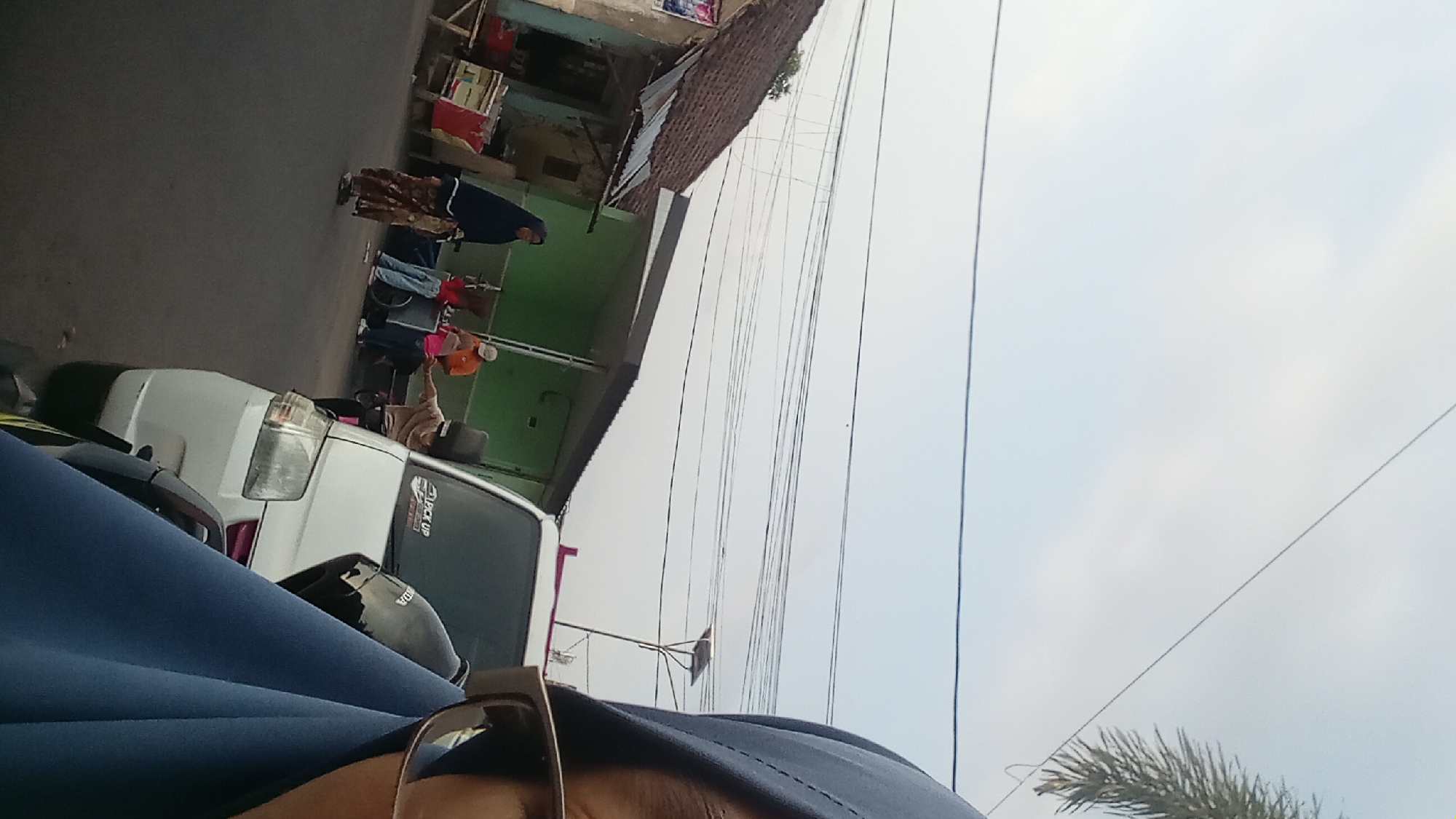 In Mataram, Indonesia, a street scene unfolds under a hazy sky, bisected by a network of overhead power lines. A person in a blue hijab, partially visible and out of focus, takes a selfie in the foreground, their glasses reflecting the scene.
To the left, a simple building with a tiled roof and weathered walls stands, suggesting a local shop or residence. In front of it, a few people are gathered around carts, possibly vendors. One woman, dressed in a dark hijab and a patterned skirt, stands near the building. Further down the street, a white van, labeled "PICK UP," is parked. A person in an orange shirt is visible near a cart, and another person in a tan shirt is leaning towards the van. The street itself appears to be paved, and the overall atmosphere suggests a casual, everyday setting in an Indonesian city.