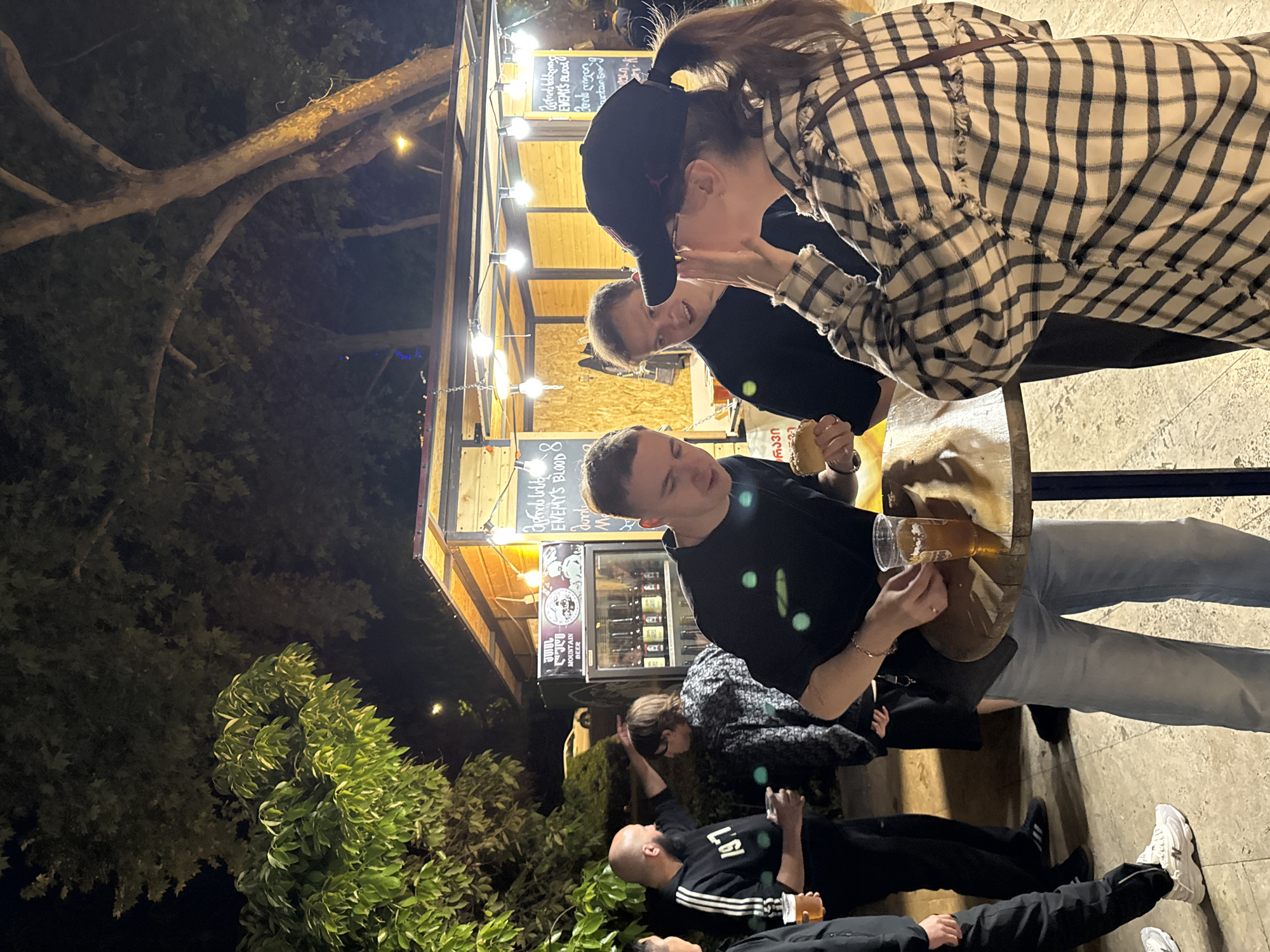 The image shows a group of people gathered around a food stall outdoors at night. The stall is illuminated by string lights and has a sign in a language that is not English. A man is holding a clear plastic cup with a liquid that appears to be beer, while another man in the background is holding a sandwich or burger. A woman wearing a plaid shirt and black baseball cap is talking to a person behind the counter. Below the counter, some people are sitting on the ground, with one man visible lying down. The environment features trees and plants in the background, suggesting a park or public space. The location is in the city of Unknown, Unknown.