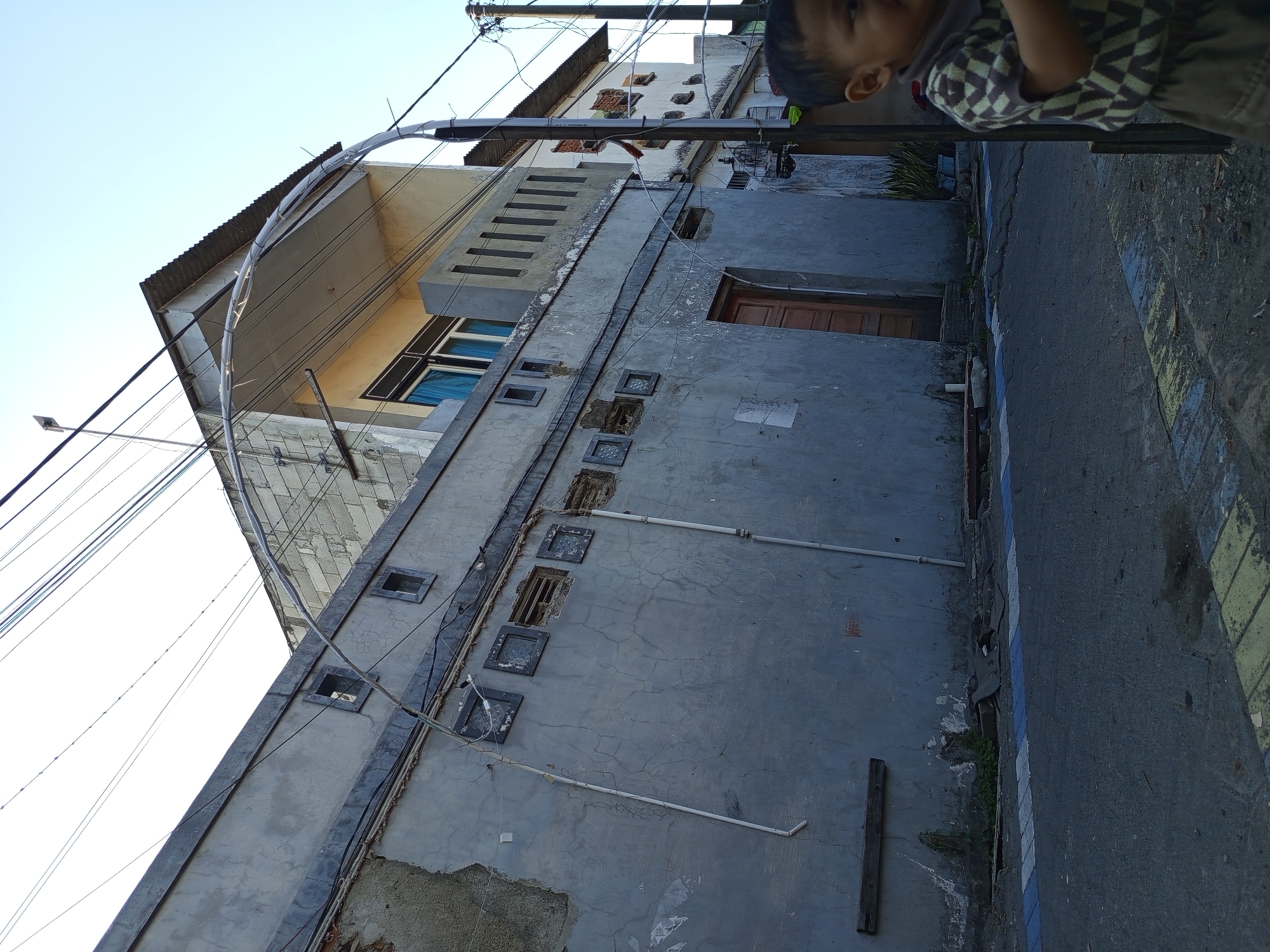 An outdoor, eye-level, medium shot shows a street scene in Tebel, Indonesia. The scene is dominated by a weathered, gray building with a dark roofline. The building has several small, square windows, some of which are broken or have grates. A brown wooden door is visible on the side of the building. Overhead, a dense network of power lines crisscrosses the sky. In the foreground, a young child, wearing a checkered shirt and brown shorts, is partially visible on the right side of the frame, looking to the right. The street is paved, with yellow and blue markings on the curb. Some greenery and debris are present along the side of the street. The sky is clear and blue.