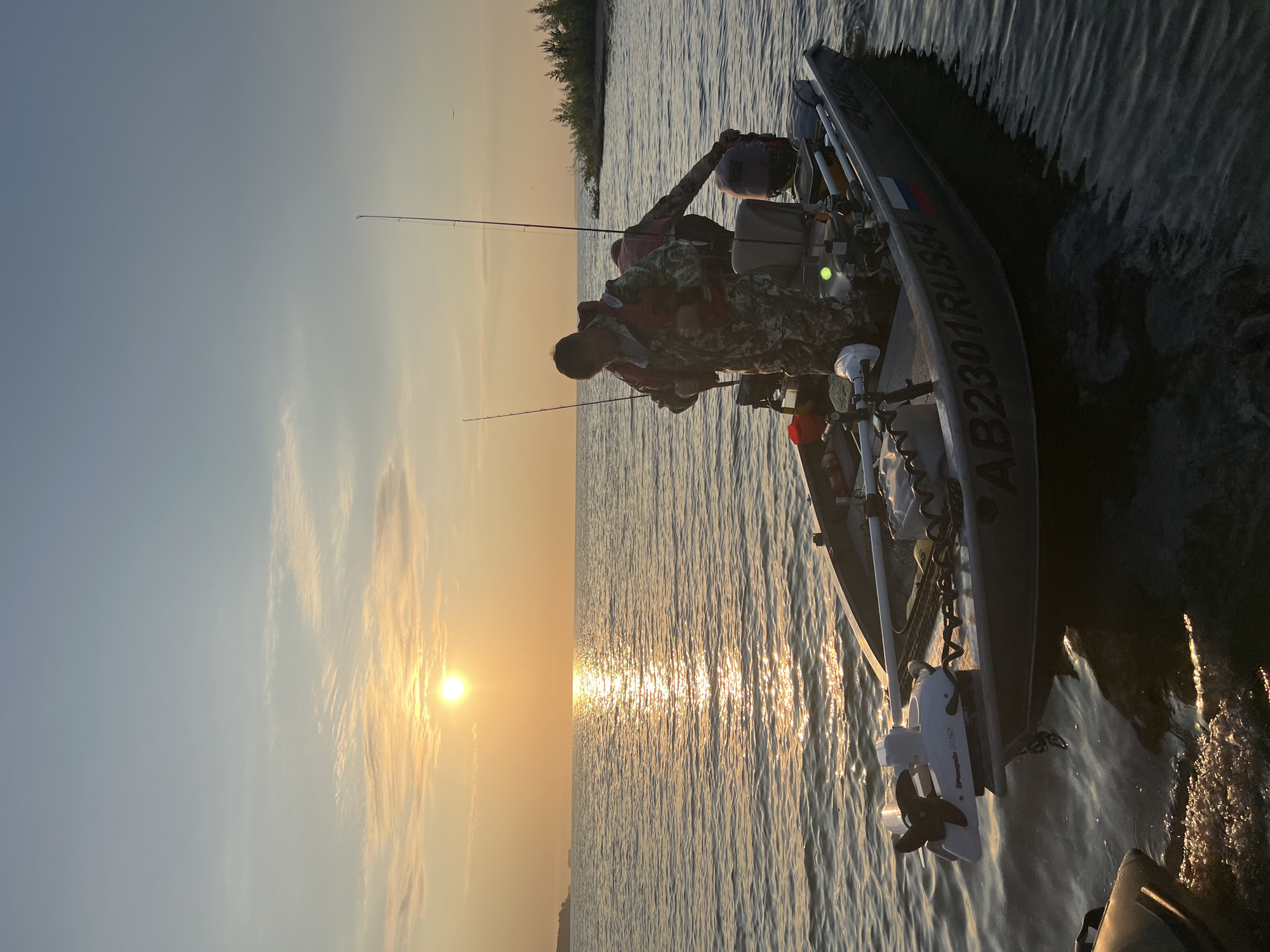 An overhead medium shot captures a man wearing a camouflage hunting suit and an orange life vest in a boat on a body of water. He is adjusting the life vest. Another person, also in camouflage attire, is partially visible behind him, their hands on the boat's motor. Two fishing rods are erected in the boat. The boat itself is a dark gray aluminum vessel with the registration number "AB2301RUS54" clearly visible on its side, along with a Russian flag emblem. A white electric trolling motor is mounted on the front of the boat. The scene is illuminated by the golden light of a setting or rising sun, which casts a shimmering reflection on the calm water. The horizon shows a distant landmass and a sliver of coastline with sparse trees. The sky is a soft gradient of blue and orange, with wispy clouds. The location is identified as Borovoye, Russia.