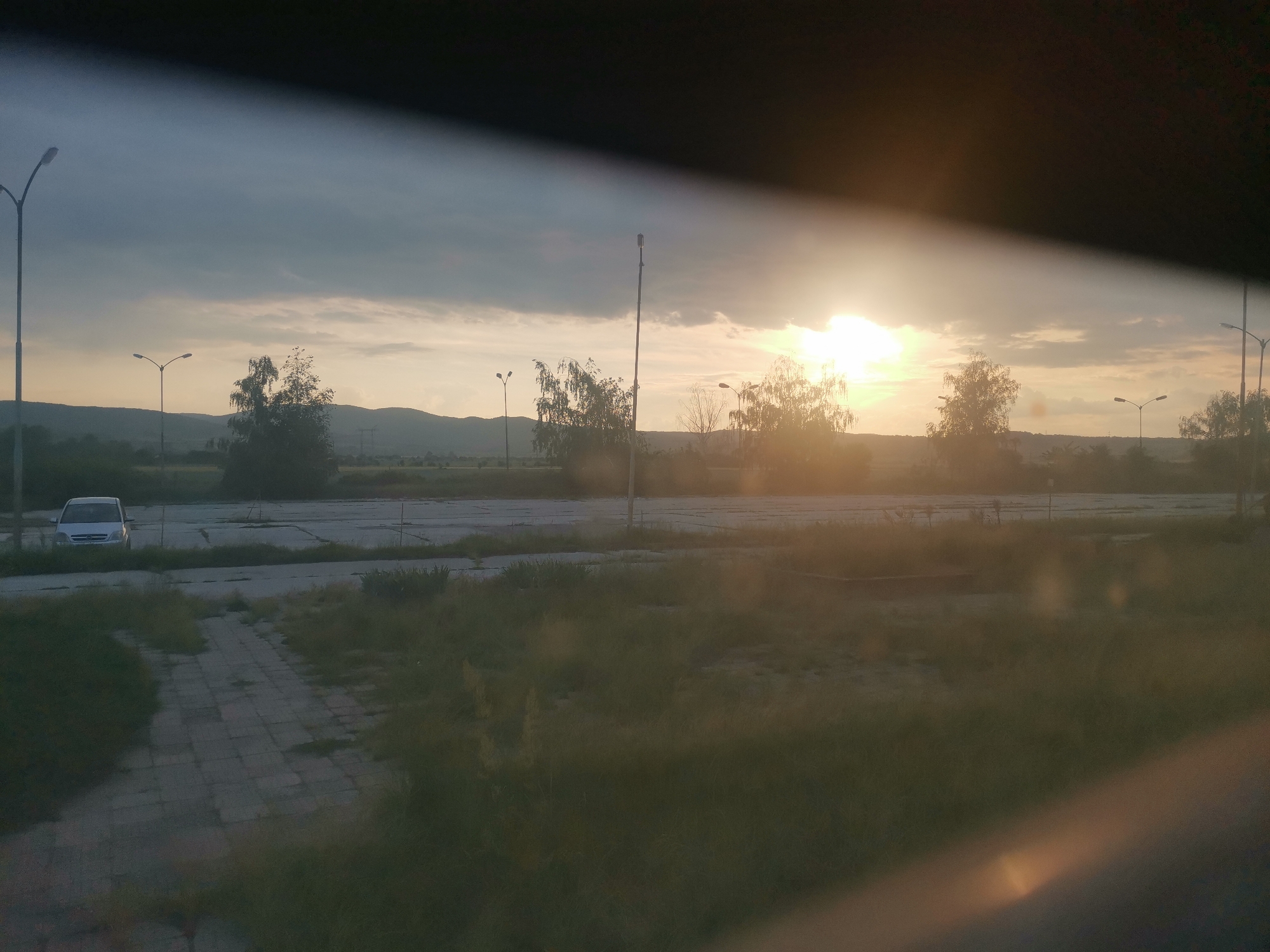 This image presents a semi-rural landscape at either sunrise or sunset in Zhivkovo, Bulgaria. The sun is low on the right side of the frame, emitting a strong glare and warm light. The sky transitions from dark grey, dense clouds at the top to lighter, warm-toned clouds near the horizon.
In the immediate foreground, a paved path composed of rectangular slabs is partially obscured by overgrown grass and weeds, which also densely covers much of the ground. Behind this, a large, cracked concrete or asphalt surface, indicative of a former parking lot or industrial area, extends across the midground. A silver minivan or SUV is parked facing left on this paved surface.
Several dark-colored streetlights with double-arm fixtures are visible throughout the midground, interspersed with deciduous trees; some trees appear to have sparse foliage. A range of hills or low mountains defines the distant horizon under the bright sky. No individuals are present in the scene. The top and bottom sections of the image feature dark, semi-transparent bands, suggesting the photograph was taken from inside a vehicle or through a window.