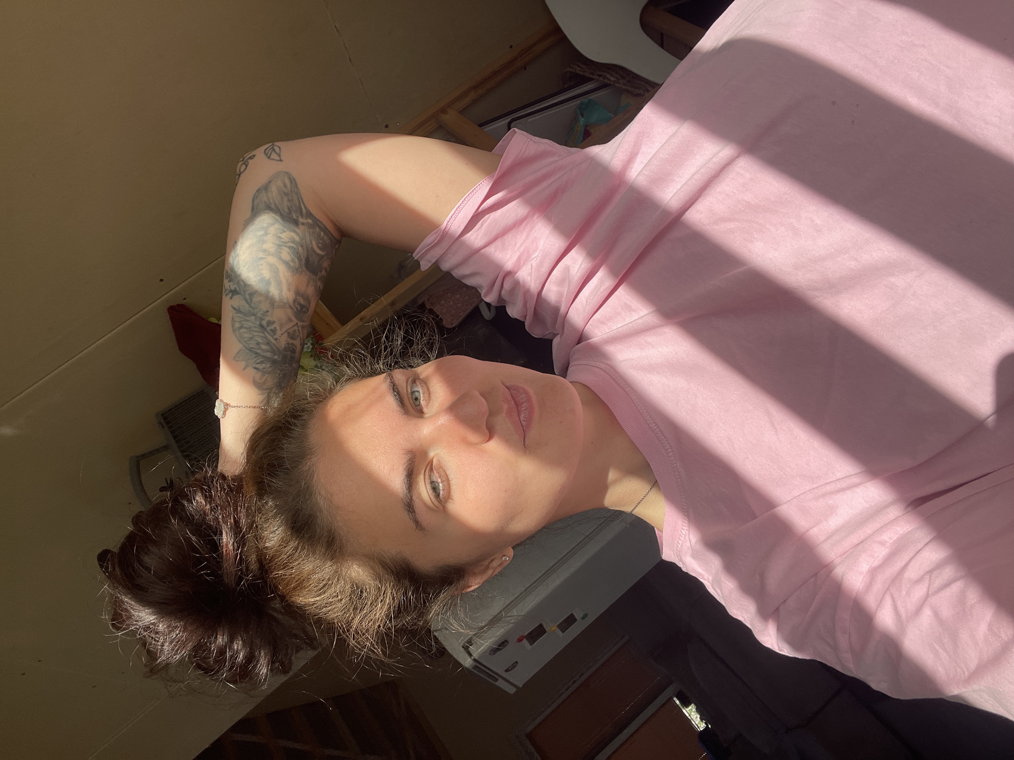 A young woman with brown hair in a messy bun is taking a selfie in what appears to be a dimly lit room. Sunlight casts strong diagonal shadows across her pink t-shirt and face. Her left arm is raised, and she's touching her hair. She has a tattoo on her left arm and a bracelet on her wrist. The background shows some furniture and a refrigerator. There are no visible elements that suggest the location is the city of Мосметаллоконструкция Онт, Russia.