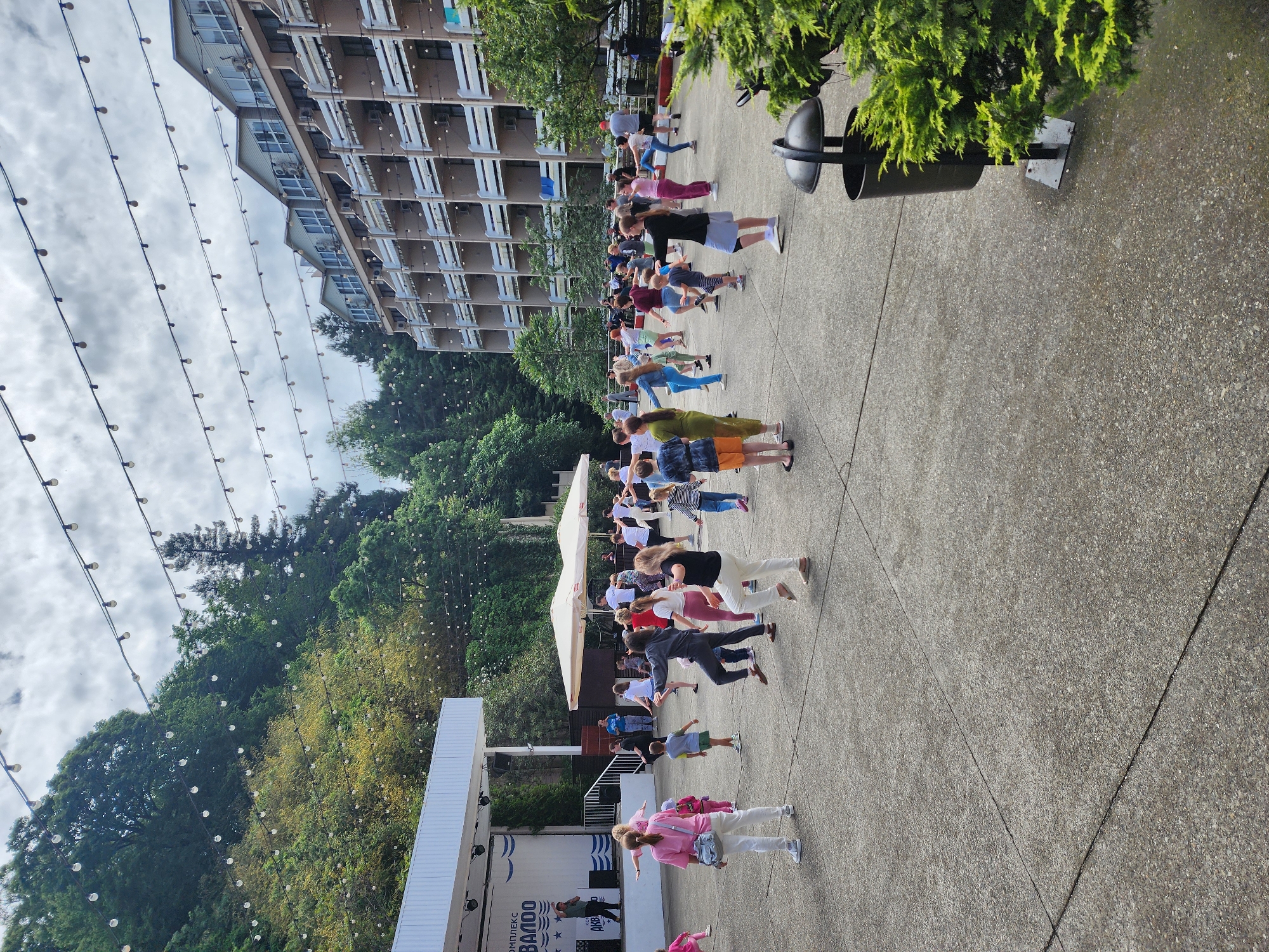 The image depicts a public outdoor event in Sochi, Russia. A group of approximately 30-40 people are participating in what appears to be a group exercise or dance activity on a paved plaza. The participants are mostly of varying ages and dressed in casual attire. In the background is a stage with a sign in Russian. A multi-story building is visible in the background to the left, along with trees and string lights hanging overhead. A trash receptacle is located to the right. The weather appears overcast.