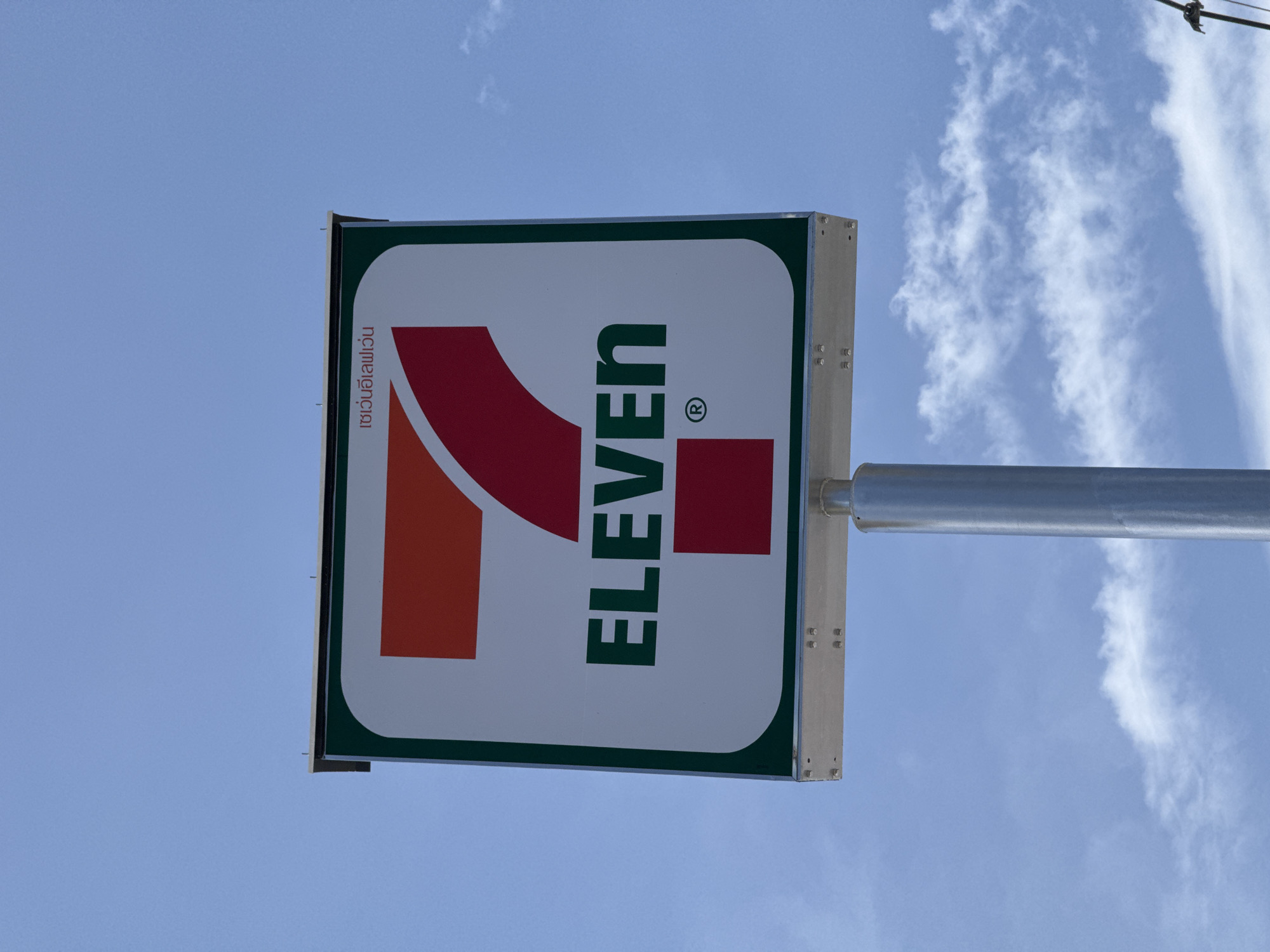 The image shows a close-up, eye-level view of a 7-Eleven sign against a blue sky with scattered clouds. The sign is square with rounded corners and a dark green border. The 7-Eleven logo, featuring the number "7" in red and orange on a white background, is prominent. The word "ELEVEN" is printed in green capital letters below the number. The Thai text "เซเว่นอีเลฟเว่น" is visible in the upper right corner of the sign. The sign is mounted on a silver metal pole. The image appears to have been taken in Ko Samui District, Thailand, given the Thai text on the sign. The lighting suggests it is daytime.