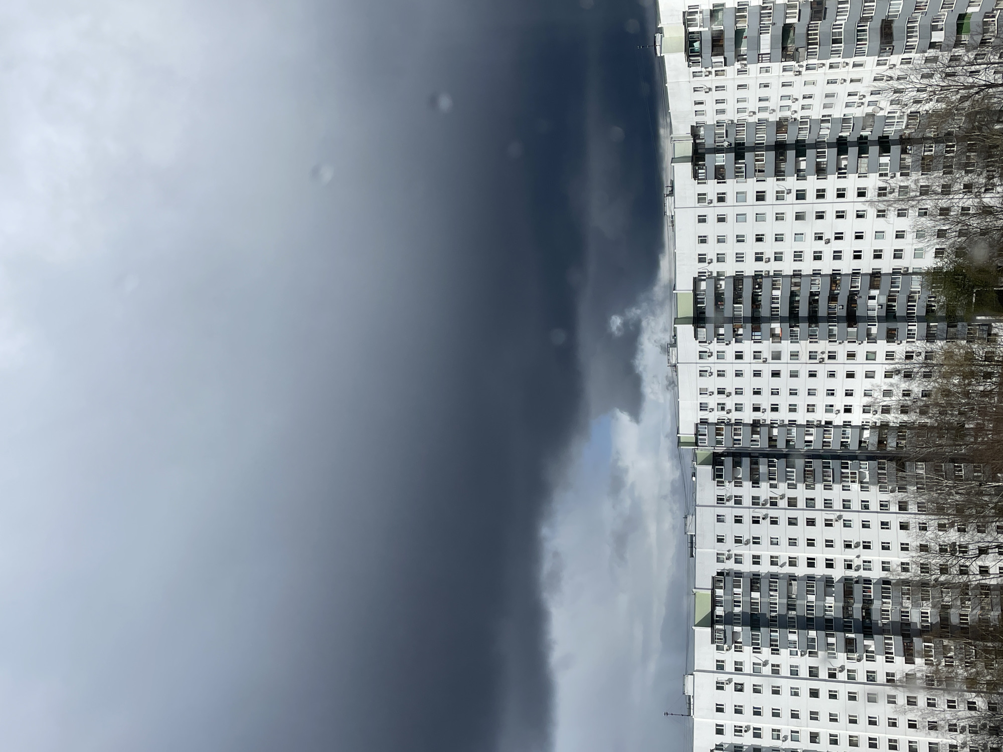 The image shows a tall, white, multi-story apartment building in Moscow, Russia, viewed against a stormy sky. The building is characterized by rows of identical windows, some with visible air conditioning units. Bare trees are visible to the right of the building. The sky is dominated by dark, ominous storm clouds that create a stark contrast with the building's facade. The presence of rain on the window indicates that it is raining.