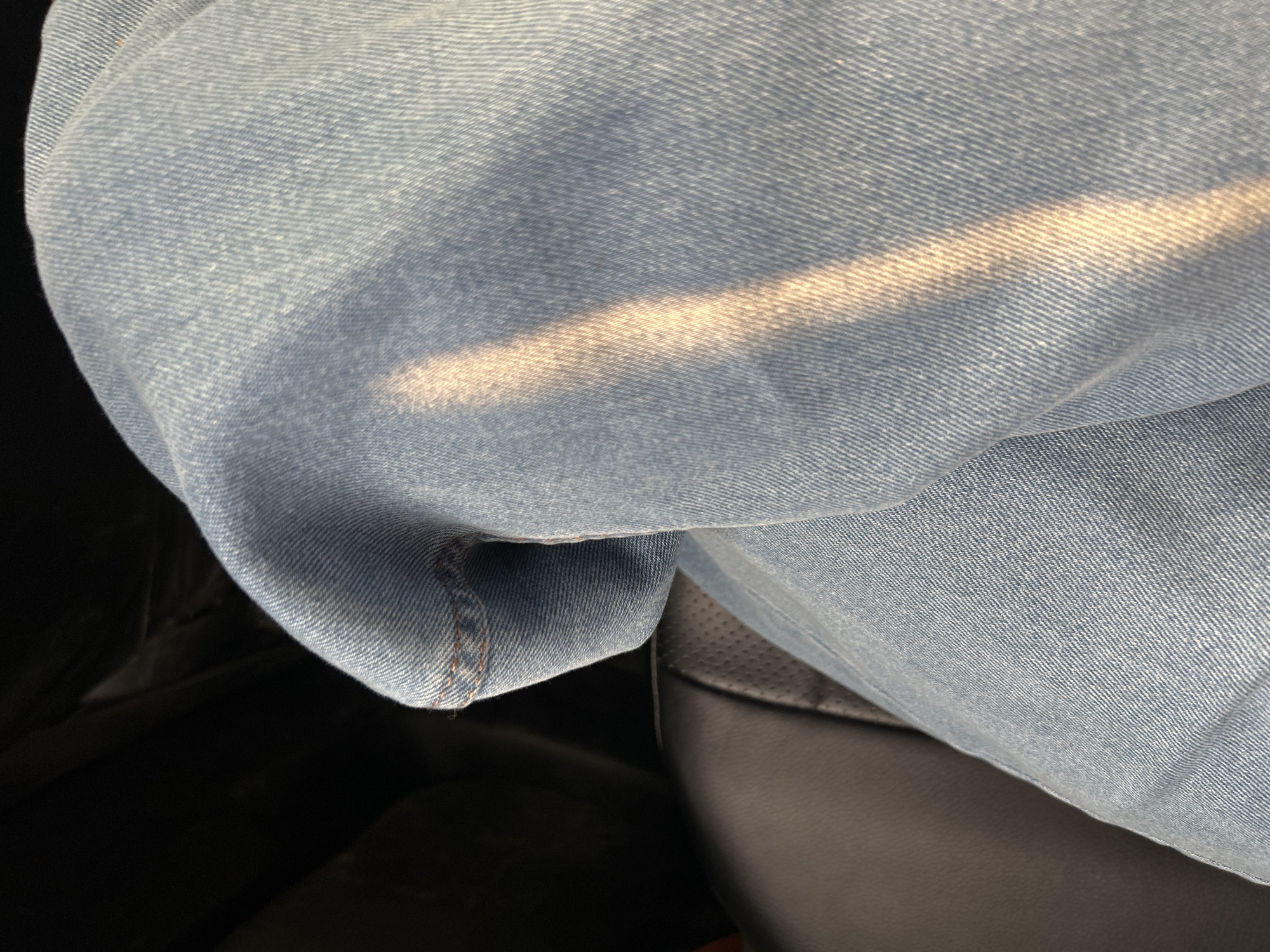 The image shows a close-up view of light-blue denim jeans. A diagonal shaft of light illuminates the fabric, highlighting the texture and weave. The jeans are creased and folded, suggesting they are being worn or are resting on a surface. In the background, a dark, textured surface, possibly leather, is visible, indicating the jeans are likely in a car or on a piece of furniture. The lighting and focus suggest an indoor or shaded outdoor environment. There is no discernible location in Ivanovo, Russia.