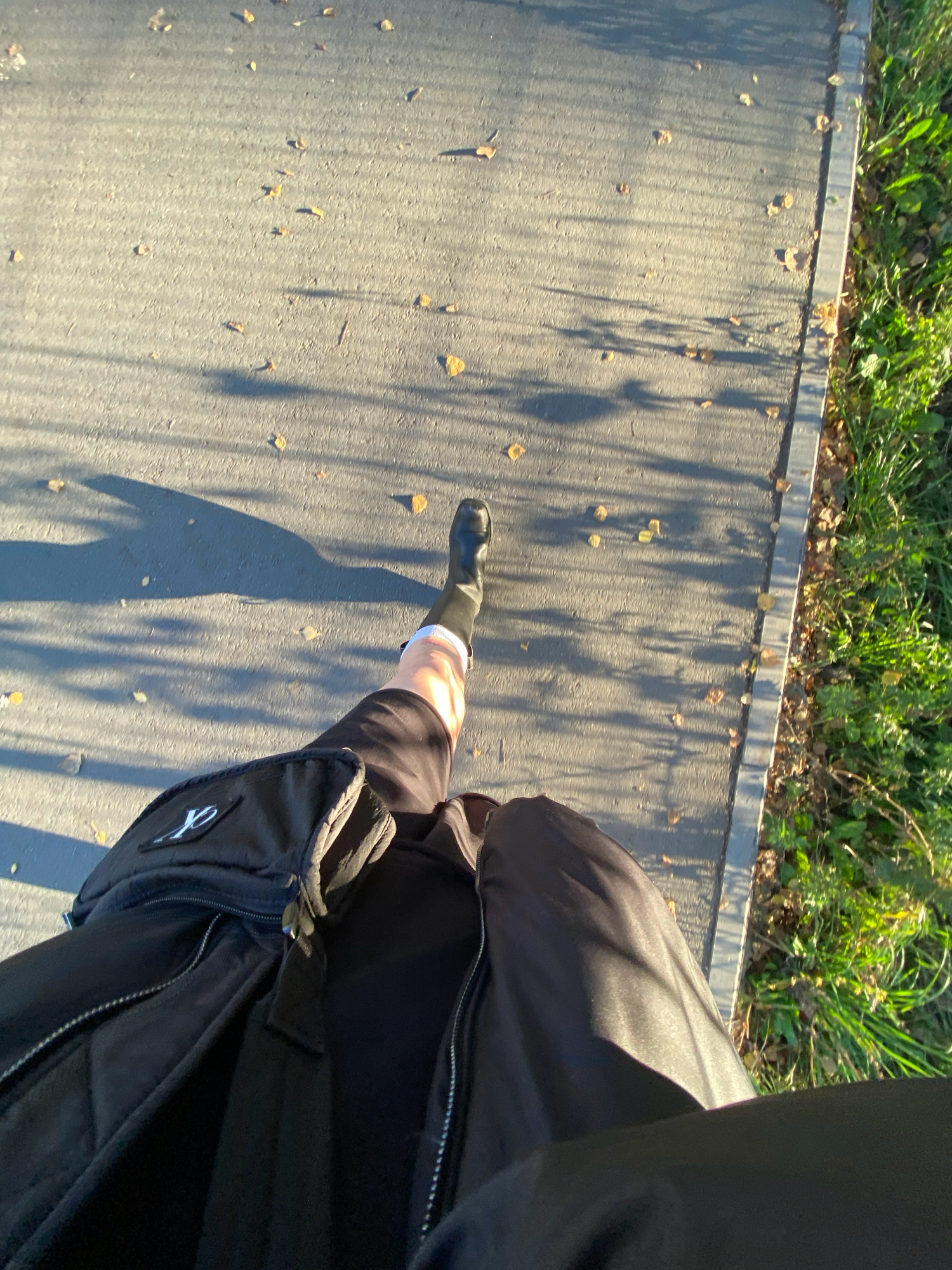 The image shows a first-person perspective of someone walking on a paved pathway with scattered autumn leaves. To the right of the pathway is a strip of green grass with some fallen leaves. The person is wearing a black boot on their right leg, which is visible from the knee down, with white socks. They are also wearing black shorts and a black jacket, with a black backpack visible on their left side. The sun is casting long shadows across the pathway. The scene is consistent with an outdoor public space in Votkinsk, Russia, during autumn.
