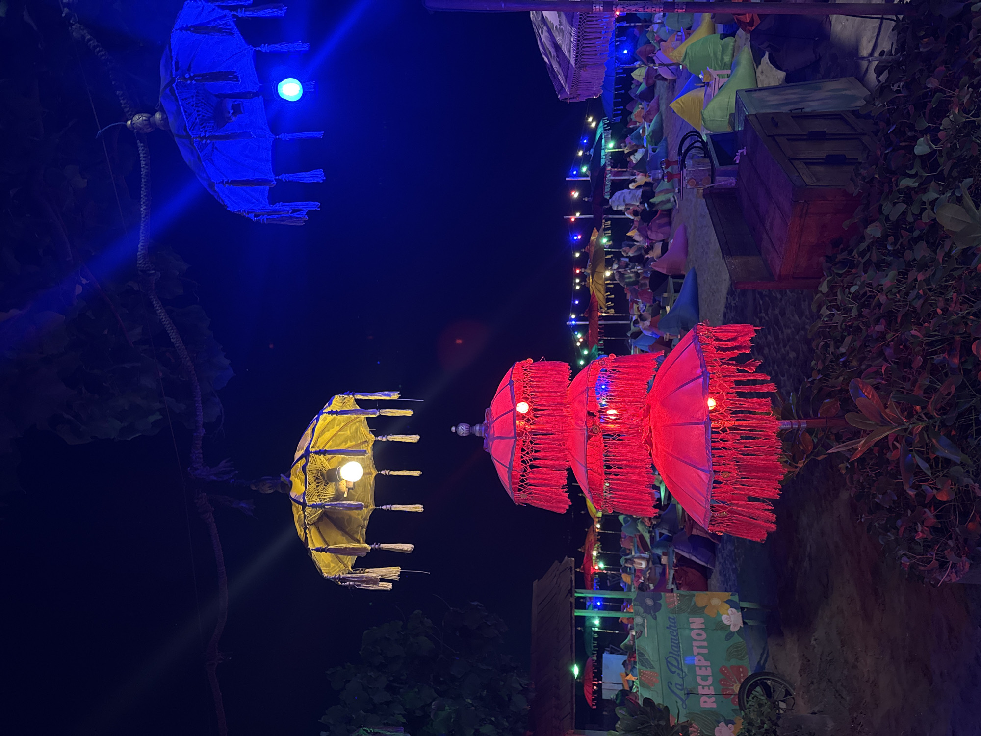The image depicts a beachside restaurant at night, likely in Kuta, Indonesia, given the vibrant atmosphere and the presence of umbrellas and beanbag seating typical of popular beach destinations. In the foreground, three large, stacked red umbrellas with decorative fringes are illuminated, casting a warm glow. To the left, a sign reads "La Plancha Reception," indicating the establishment. Beyond this, a row of colorful umbrellas, including red, yellow, and green, are set up on the sandy beach. Numerous beanbag chairs are scattered around, occupied by a crowd of people socializing and enjoying the evening. String lights are visible in the background, adding to the festive ambiance. Overhead, a yellow umbrella with tassels and a blue umbrella are suspended, further enhancing the decorative lighting. The sky is dark, suggesting it is nighttime. The scene conveys a lively and relaxed atmosphere, characteristic of a beach resort in a tropical location.