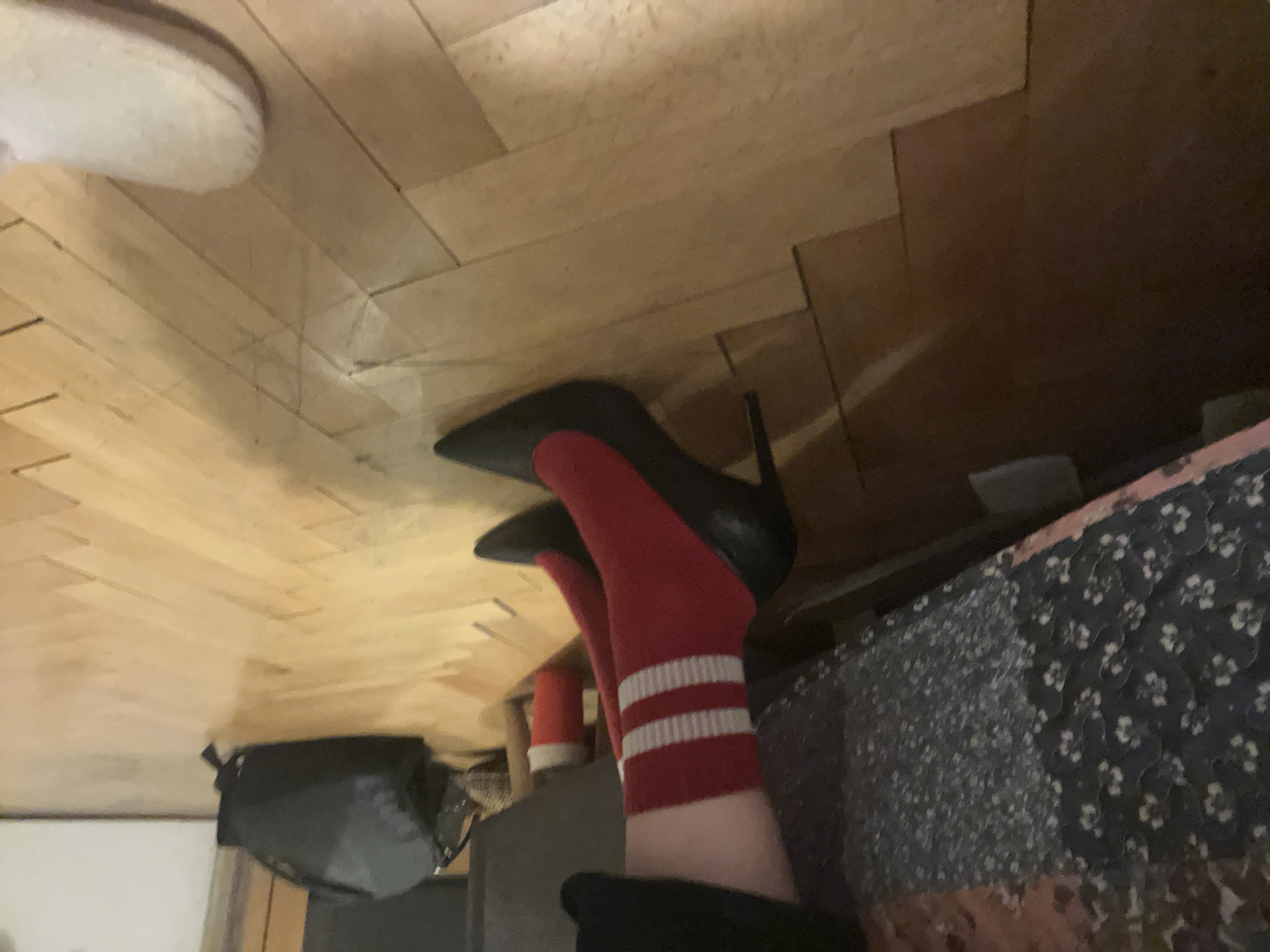The image depicts a low-angle shot of a person's feet wearing red socks with white stripes and black stiletto heels. The person is situated in what appears to be an indoor environment. The background includes a wooden floor with visible planks and a patterned object, possibly a blanket or bedspread. A gray object with a strap can be seen as well, could be a small bag. The perspective and objects present don't provide sufficient information to determine the precise location in Sofia, Bulgaria. There is no specific information to pinpoint this exact location.