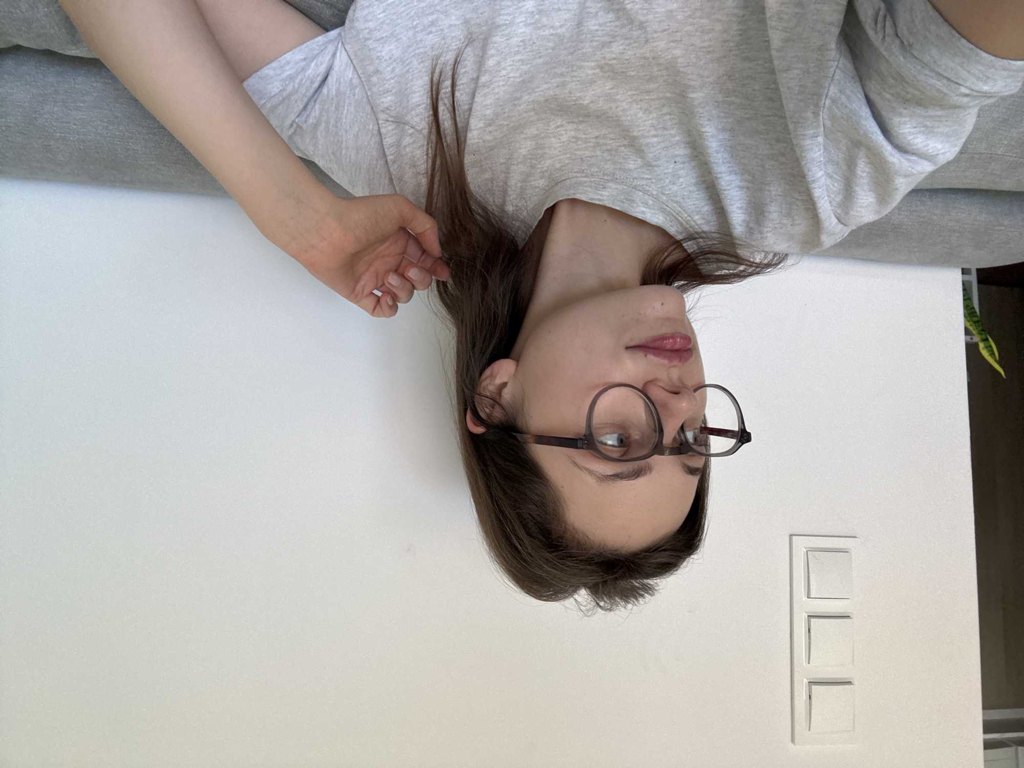 This is a vertical, slightly tilted, close-up photograph of a young person wearing glasses and a light grey t-shirt. The person's head is turned away from the camera, and their face is partially visible in profile. They have brown hair that is falling forward. Their left arm is bent and resting on their lap, with their hand gently touching their hair. The background is a plain white wall. To the right of the person, there is a white light switch with three toggles. The scene appears to be indoors, in natural light. There is no visible text in the image. The image was taken in Szklarska Poręba, Poland.