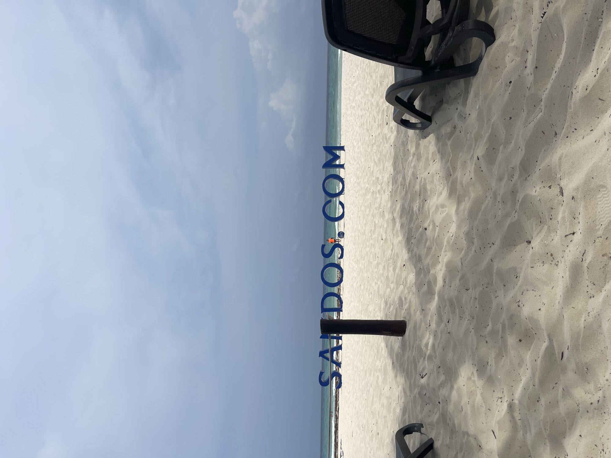 The scene is a beach in Playa del Carmen, Mexico. It shows the sand, the ocean, and the sky on a sunny day. There's a black beach chair partially visible on the right. A small, orange clad figure is visible near the water. Large, blue letters reading "SANDOS.COM" appear vertically in the image, obscuring parts of the scene. A dark brown wooden pole is partially buried in the sand.