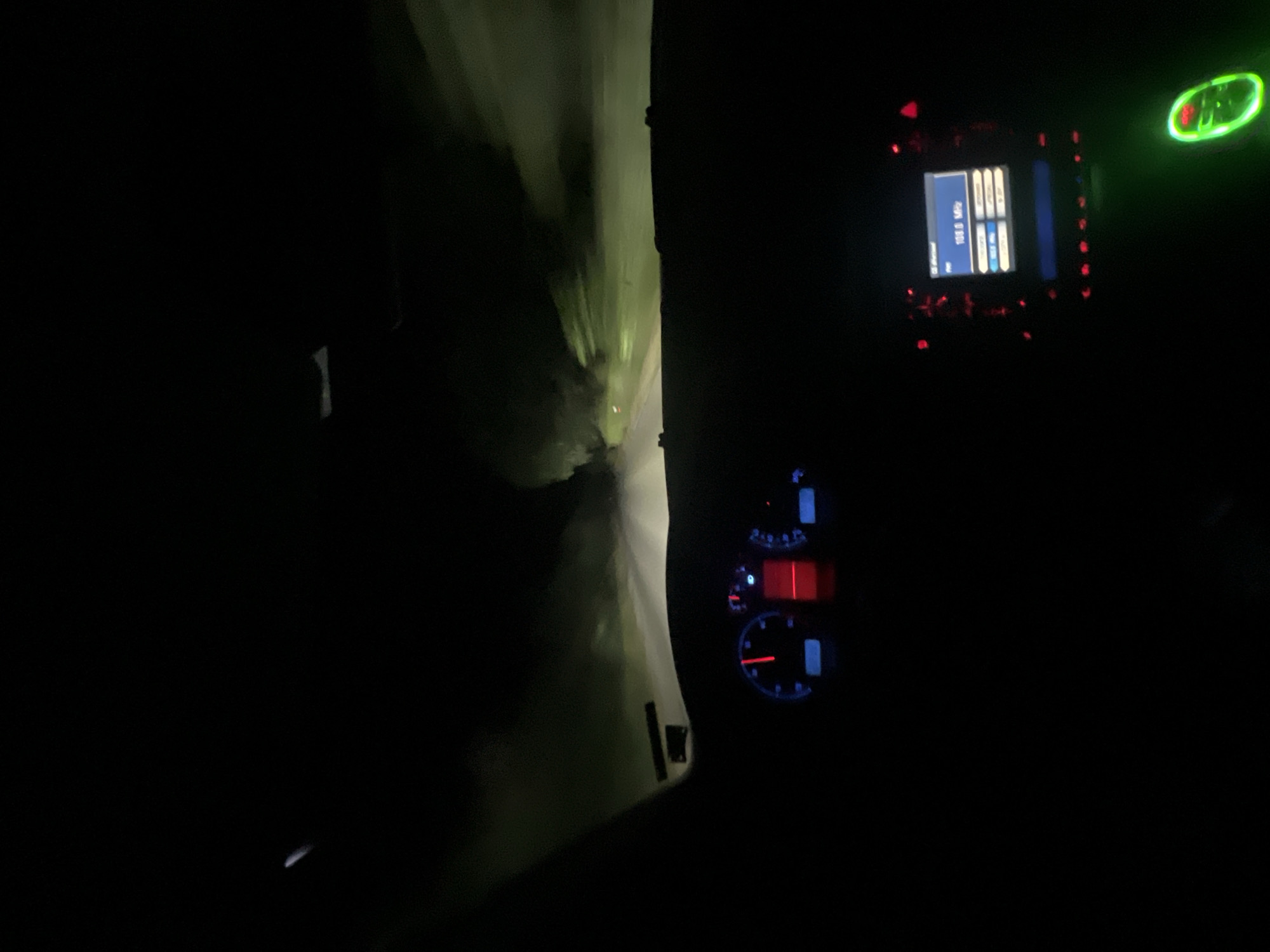 The image depicts the interior of a car driving at night, presumably on a road near Svetlina, Bulgaria. The dashboard lights are illuminated, showing a speedometer with a red needle pointing to approximately 60 km/h, and a radio display showing "108.0 MHz". The exterior view through the windshield is dark, with faint headlights illuminating a misty, tree-lined road ahead. The atmosphere suggests driving in fog or a similar atmospheric condition. There are no people or animals visible in the frame.