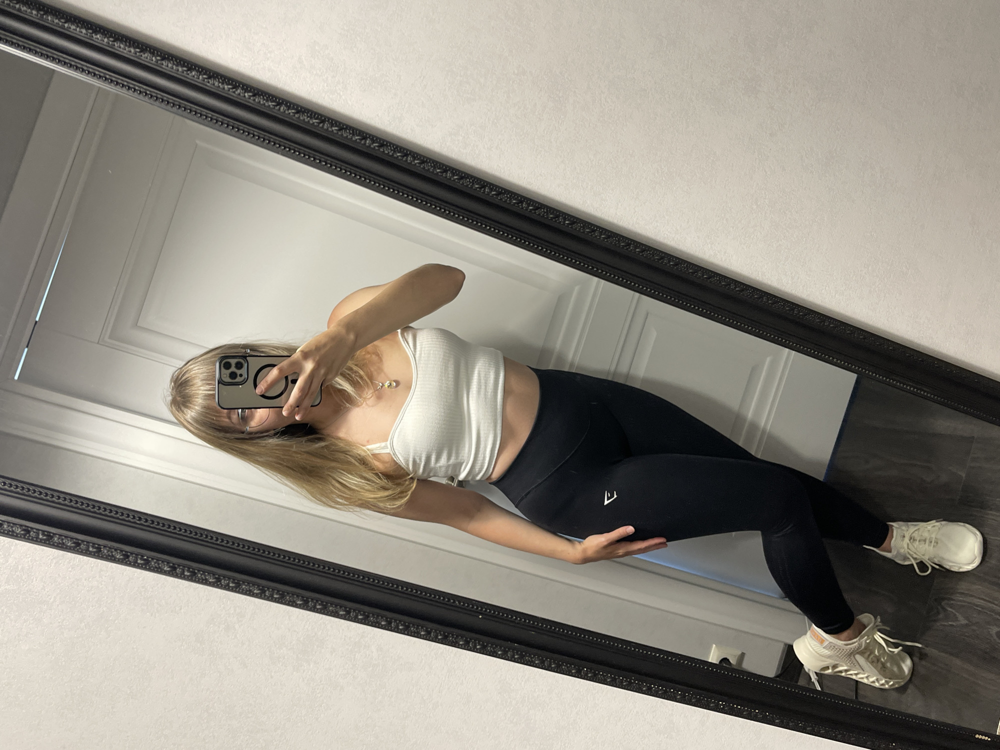 A woman is taking a mirror selfie in what appears to be an indoor, residential setting. She is standing and angled towards a large, ornate black mirror that reflects her image. She has blonde hair, wears glasses, and is dressed in a white crop top and black leggings with a white logo on the left thigh. Her right hand is resting on her hip, and she is holding a smartphone in her left hand to take the photo. She is wearing white sneakers. The background visible in the mirror includes a white door and a textured white wall. The floor is dark wood. This scene is not identifiable as being in Zhytomyr, Ukraine.