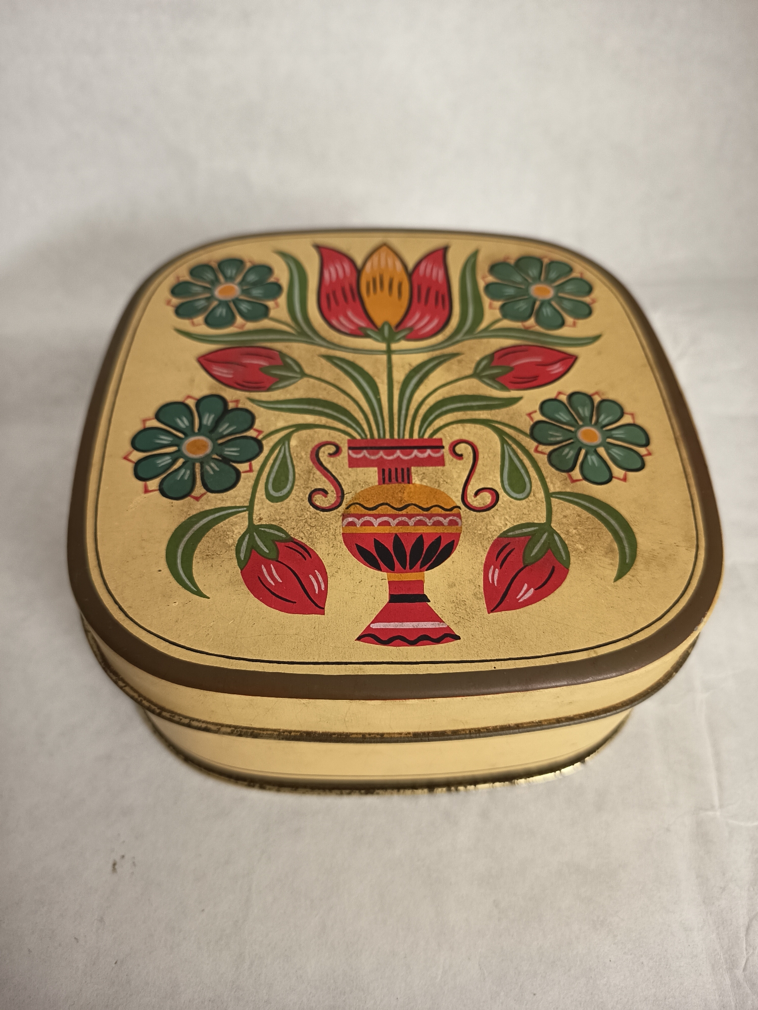 The image shows a decorative tin box with a floral design. The box is rectangular with rounded corners and has a gold-colored background. The design features a vase with flowers in red, orange, green, and blue. The vase is centrally located, and the flowers are arranged symmetrically around it. A brown trim runs along the edge of the lid. The background is a plain white surface. The box is likely of vintage or antique origin. There is no evidence that this image is connected to Wangen bei Olten, Switzerland.