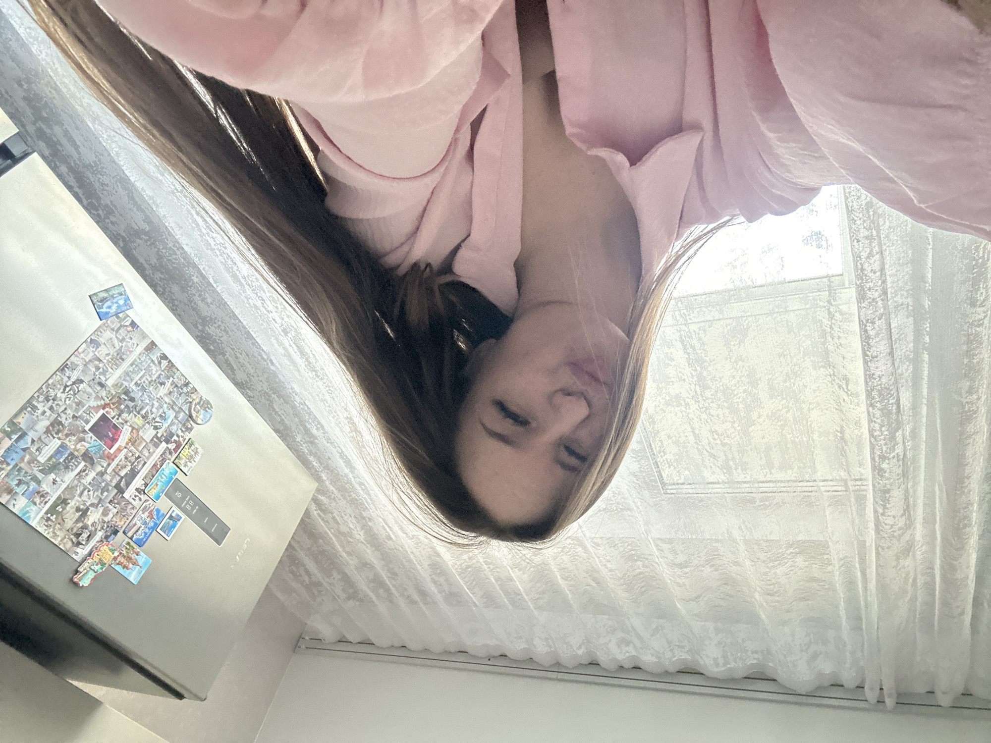 An upside-down, low-angle shot captures a young woman looking directly into the camera. She has long brown hair and is wearing a soft pink garment, possibly a robe or top. Her face is in the upper center of the frame, with a relaxed expression, one eye slightly squinted.
To the left side of the image (which would be the right if rotated), a stainless steel refrigerator is visible. It is extensively decorated with a large collage of numerous small photographs and several magnets of varying shapes and sizes, including rectangular ones and a colorful, irregularly shaped magnet. A black control panel or display on the refrigerator shows some faint white text and symbols.
To the right side (which would be the left if rotated), a window is covered by white, sheer curtains that are slightly textured. Bright, diffused natural light streams through the window, indicating it is daytime. The overall setting is an indoor environment, likely a kitchen or living area, suggested by the presence of the refrigerator and window with curtains. The unusual perspective lends a candid or playful atmosphere to the scene.