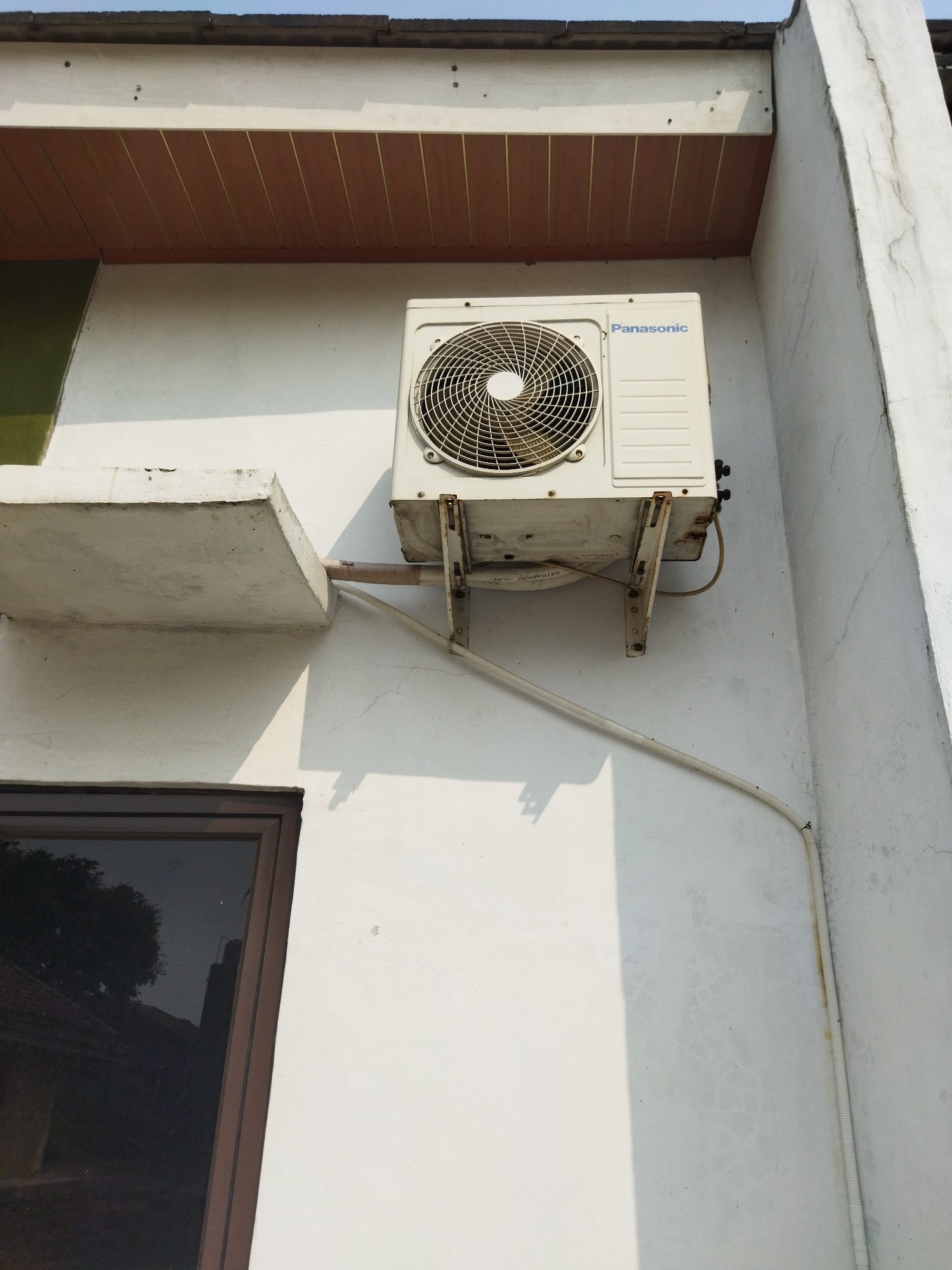 The image shows an outdoor unit of a Panasonic air conditioner mounted on the white wall of a building. The unit is situated below a wooden soffit that is part of the building's roofline. A concrete ledge extends from the wall to the left of the air conditioner. A dark-tinted window with a brown frame is visible below the concrete ledge. The air conditioner is connected to a pipe that runs down the wall, secured with black zip ties. The scene is lit by sunlight, casting shadows on the wall. The overall environment appears to be an exterior wall of a residential or commercial building, likely in a tropical or subtropical climate given the presence of the air conditioner and the bright sunlight. The location is identified as Suka Sari, Indonesia. There are no people or animals present in the image, and no specific actions or interactions are depicted. The focus is on the architectural elements and the air conditioning unit.
