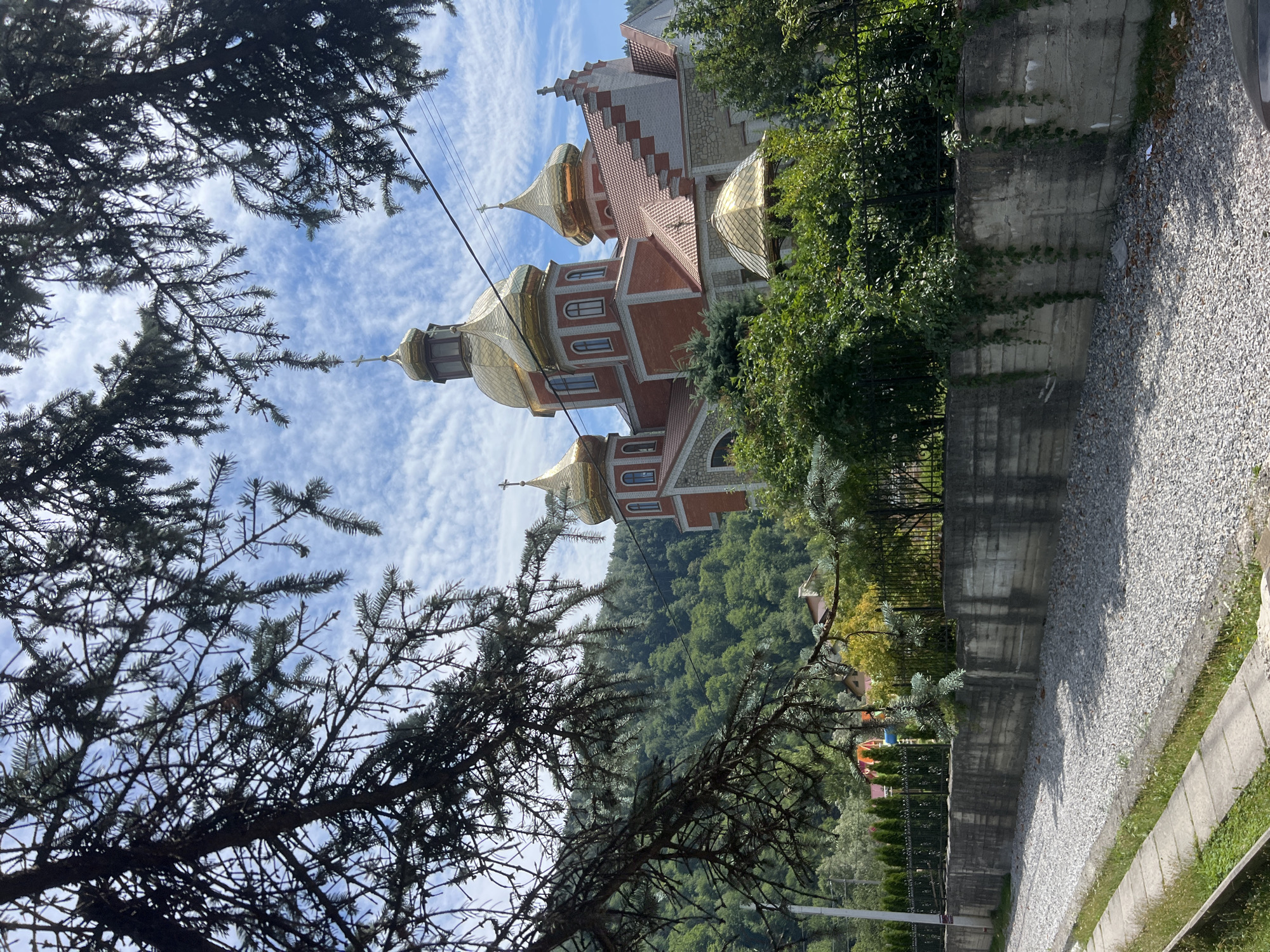An outdoor, low-angle shot captures the facade of a red brick church with golden domes, set against a backdrop of lush green mountains and a bright blue sky dotted with white clouds. The church features multiple domes, the largest of which is centrally located, topped with a cross. The architecture includes stone accents and a red tiled roof. In the foreground, a concrete wall runs horizontally across the image, bordering a gravel-covered area and a narrow concrete path with grass. Evergreen trees and branches of a conifer tree frame the scene from the left and top. The surrounding environment suggests a semi-rural or mountainous location, consistent with Yaremche, Ukraine. There are no people visible in the image. The lighting indicates it is daytime.
