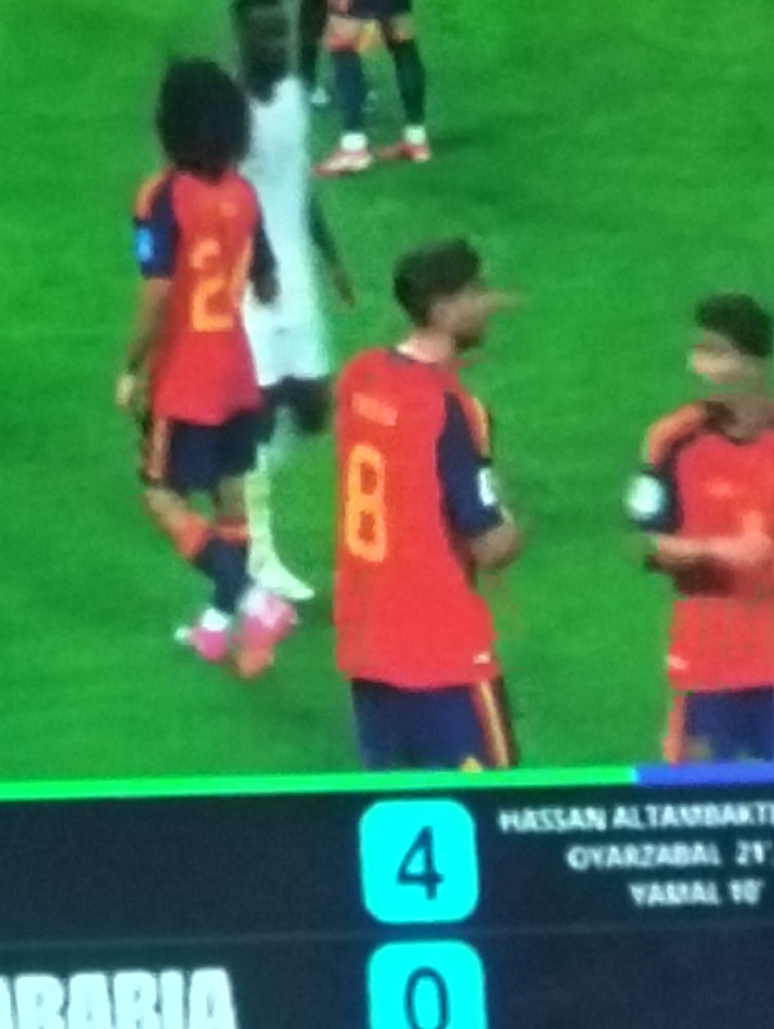 The image depicts a soccer match with a scoreboard overlay. On the field, several soccer players in red and blue uniforms are visible. One player in the foreground is wearing a red jersey with the number 2 on the back and dark shorts. Another player in a red jersey with the number 8 is also visible. The scoreboard displays "4" in a cyan box on the left and player names with times on the right, including "HASSAN ALTAMBAKTI", "OYARZABAL 21'", and "YAMAL 10'". The field is green, indicating a soccer pitch. The text "ARABIA" is partially visible at the bottom left of the scoreboard. The image is pixelated, making fine details difficult to discern. The exact location of Payagaboh, Indonesia, is not depicted in the image.