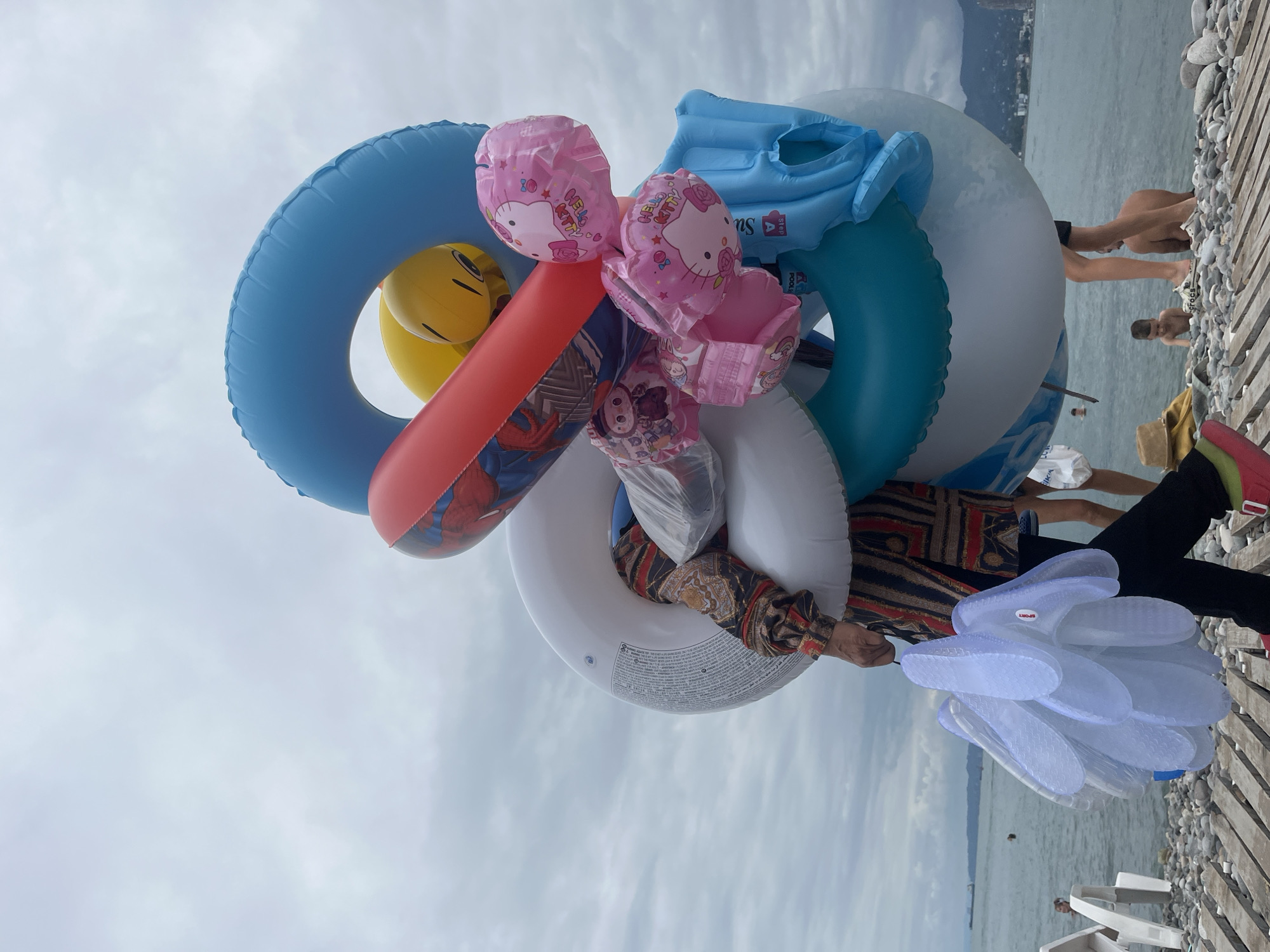 An overcast day at the pebble beach in Batumi, Georgia, where a person is holding a large stack of inflatable items, including swim rings and balloons. The person is wearing a black outfit with a vibrantly patterned jacket and bright pink shoes. Among the inflatables are blue and white swim rings, a red ring with a superhero design, yellow character balloons, and pink Hello Kitty balloons. To the left, a bunch of clear plastic flip-flops are also being carried. In the background, the sea stretches to the horizon, with a few people swimming or wading. Further inland, mountains are visible beneath the cloudy sky. The foreground features a wooden boardwalk and scattered pebbles.