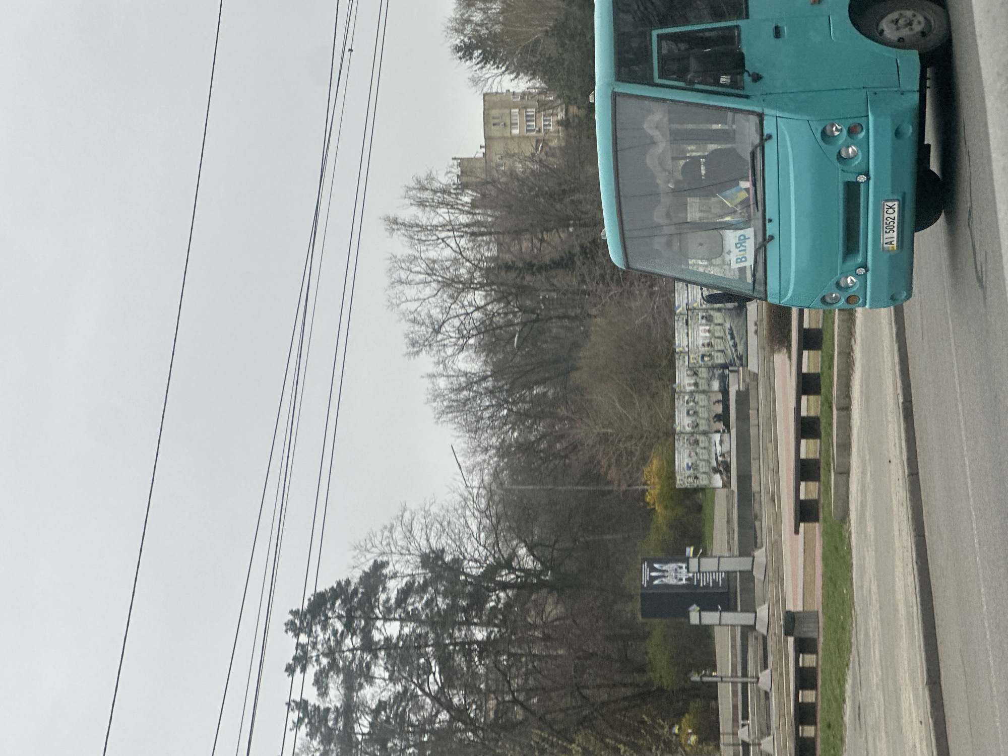 The image shows an urban scene in Kyiv, Ukraine. In the foreground, a turquoise bus is parked on the side of a road, facing right. The license plate reads "AI 5052 CK." To the left of the bus, there are several grey concrete structures or posts with dark stripes, and behind them is a billboard with black-and-white imagery. Deciduous trees with bare branches are also visible. In the background, a multi-story building stands amidst the trees. The sky is overcast and grey. Overhead electrical cables stretch across the upper left portion of the image.