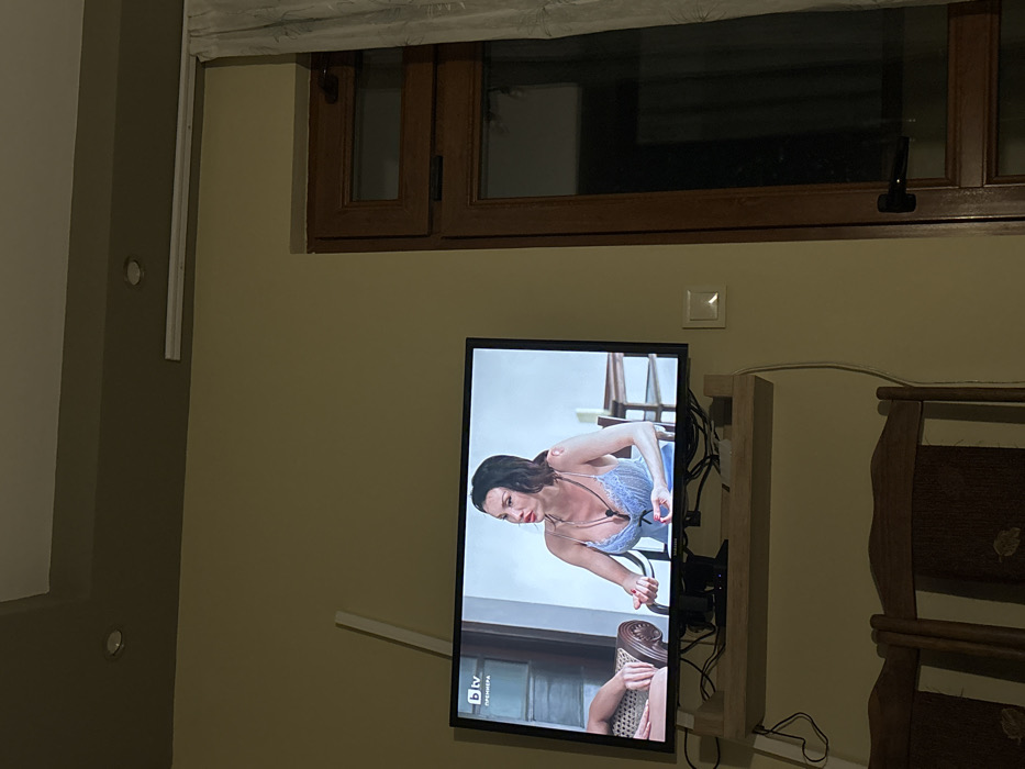 The image depicts an indoor scene, specifically a room located in Burgas, Bulgaria. The room has light olive-green walls and a recessed ceiling section with dark brown trim and two embedded circular spotlights. A white linear light fixture is also visible on the ceiling.
A black flat-screen television is centrally mounted on a light-colored wooden shelf, positioned against the left wall. Visible beneath the television are a collection of black cables and wires. The television displays a program from the Bulgarian channel "bTV", indicated by the logo and the word "ПРЕМИЕРА" (PREMIERE) in the upper left corner of the screen. The program features a woman with dark hair, wearing a light blue, low-cut, camisole-style top with thin straps and multiple necklaces. She is seated, leaning forward, with her hands clasped or gesturing, appearing to be engaged in conversation. A person's arm and a wicker chair are partially visible to her left on the screen, and blurred background elements are to her right.
To the right of the television, a dark brown wooden-framed window or glass door is visible, partially obscured by a light-colored curtain hanging on a track. The outside view through the window is dark. A white rectangular wall-mounted light switch is located on the wall between the television and the window/door. Below the wooden shelf, the tops of two wooden chairs with patterned dark fabric seats are partially visible. The room is moderately lit, with illumination primarily from the television screen and ceiling fixtures.