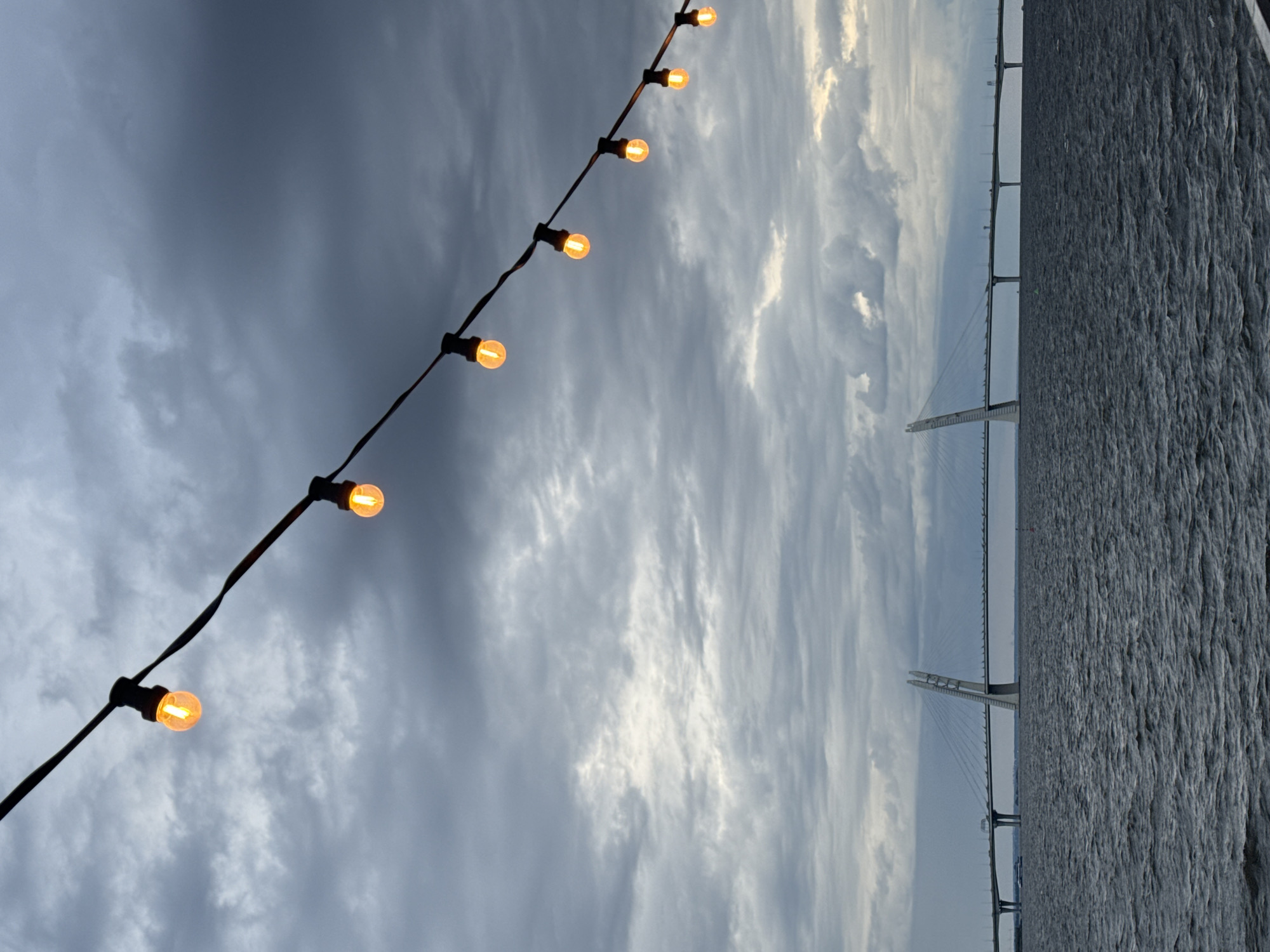 This is a medium shot photograph of a body of water, a large bridge, and a string of lights against a cloudy sky. The water in the foreground is dark and choppy, with visible ripples. In the middle ground, a long, modern cable-stayed bridge stretches across the water. The bridge has distinctive, tall, A-shaped pylons. A single boat is visible on the water to the left of the bridge. In the upper portion of the image, a string of illuminated, amber-colored lights hangs diagonally from the top left to the bottom right. The sky is overcast with heavy, grey clouds. The scene is likely Saint Petersburg, Russia, given the architecture of the bridge, which appears to be the Western High-Speed Diameter.
