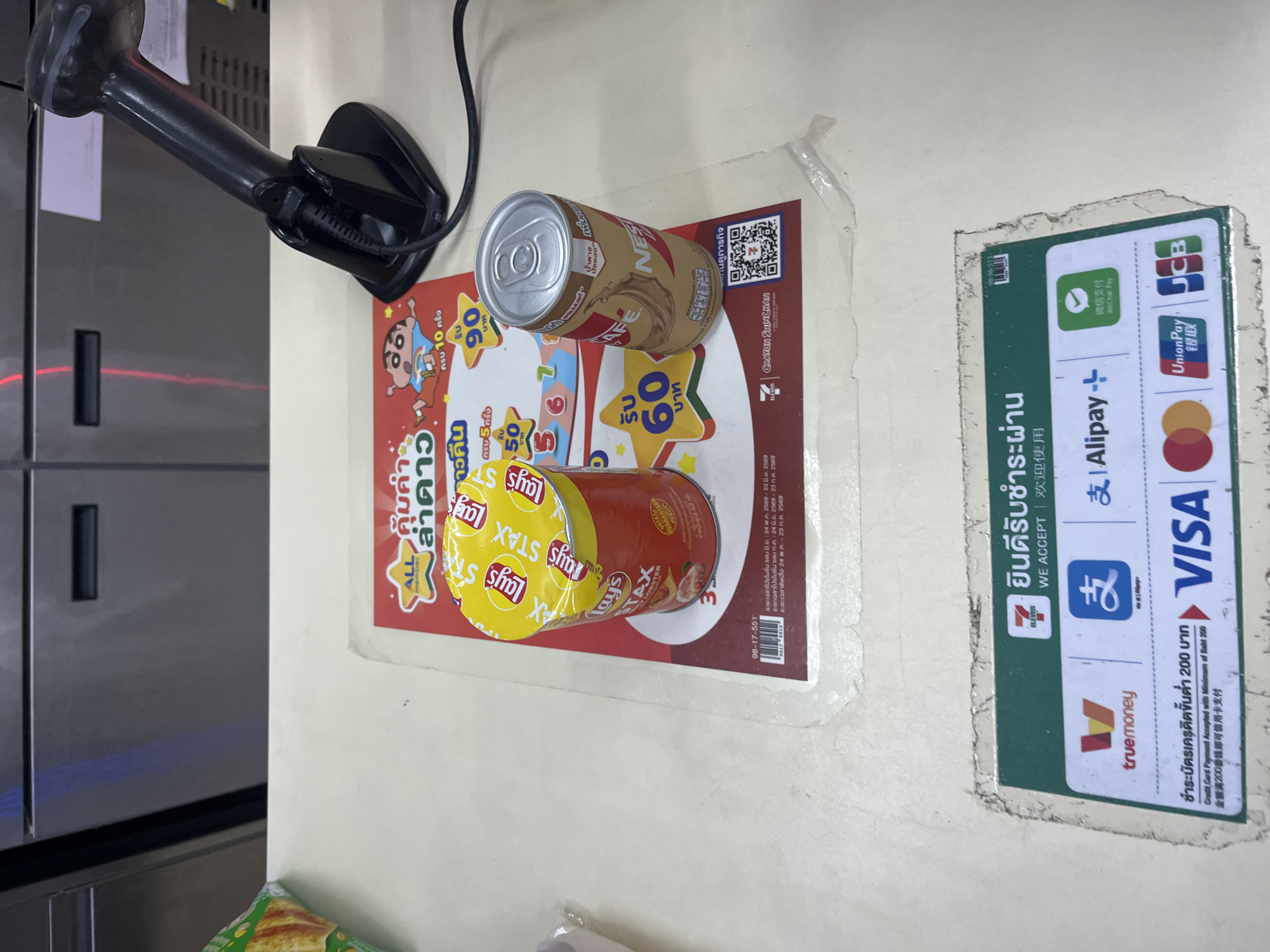 The image shows a close-up of a counter in a retail establishment, likely a convenience store in Rawai, Thailand, judging by the Thai language on the payment acceptance signage. On the counter, a red and yellow cylindrical can of Lay's Stax potato chips and a can of Nestle coffee are prominently displayed on a red promotional flyer. The flyer features cartoon characters, including Crayon Shin-chan, and advertises promotions, with prices like "60 Baht" and "90 Baht" visible. A barcode scanner with a red laser beam is positioned behind the products. Below the promotional flyer, a green and white sticker displays various payment acceptance logos, including TrueMoney, Alipay, WeChat Pay, Visa, Mastercard, UnionPay, and JCB, indicating that these are accepted payment methods. The overall environment appears to be a service counter in a store, suggesting a point-of-sale transaction is either about to occur or has just concluded.