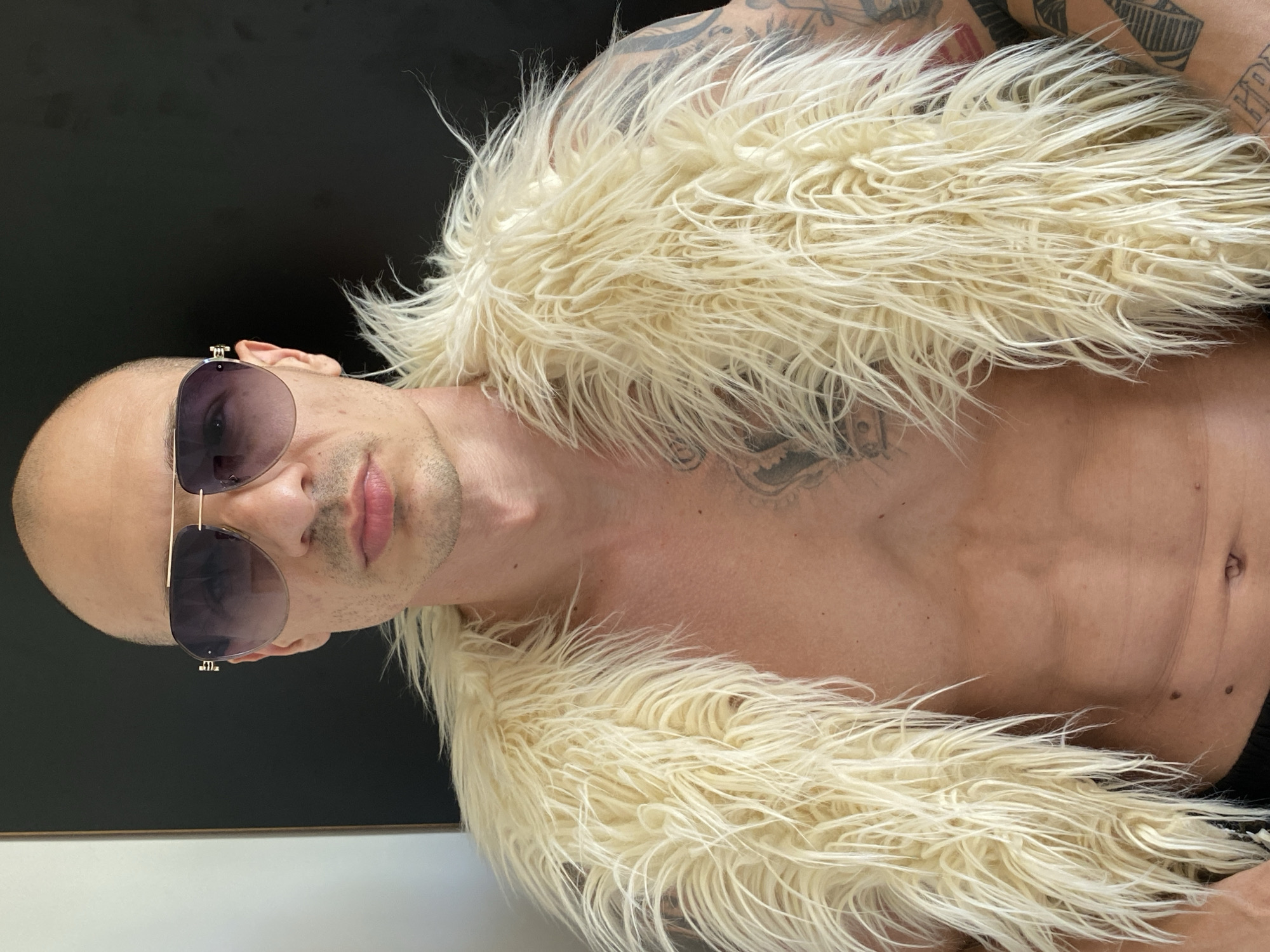 A shirtless, bald man wearing sunglasses and a cream-colored, shaggy vest is lying down. The background consists of a black wall, which is partially obscured by the man's shoulder. The man has visible tattoos on his chest and arms. The location in Pleven, Bulgaria, is not discernible from the image.