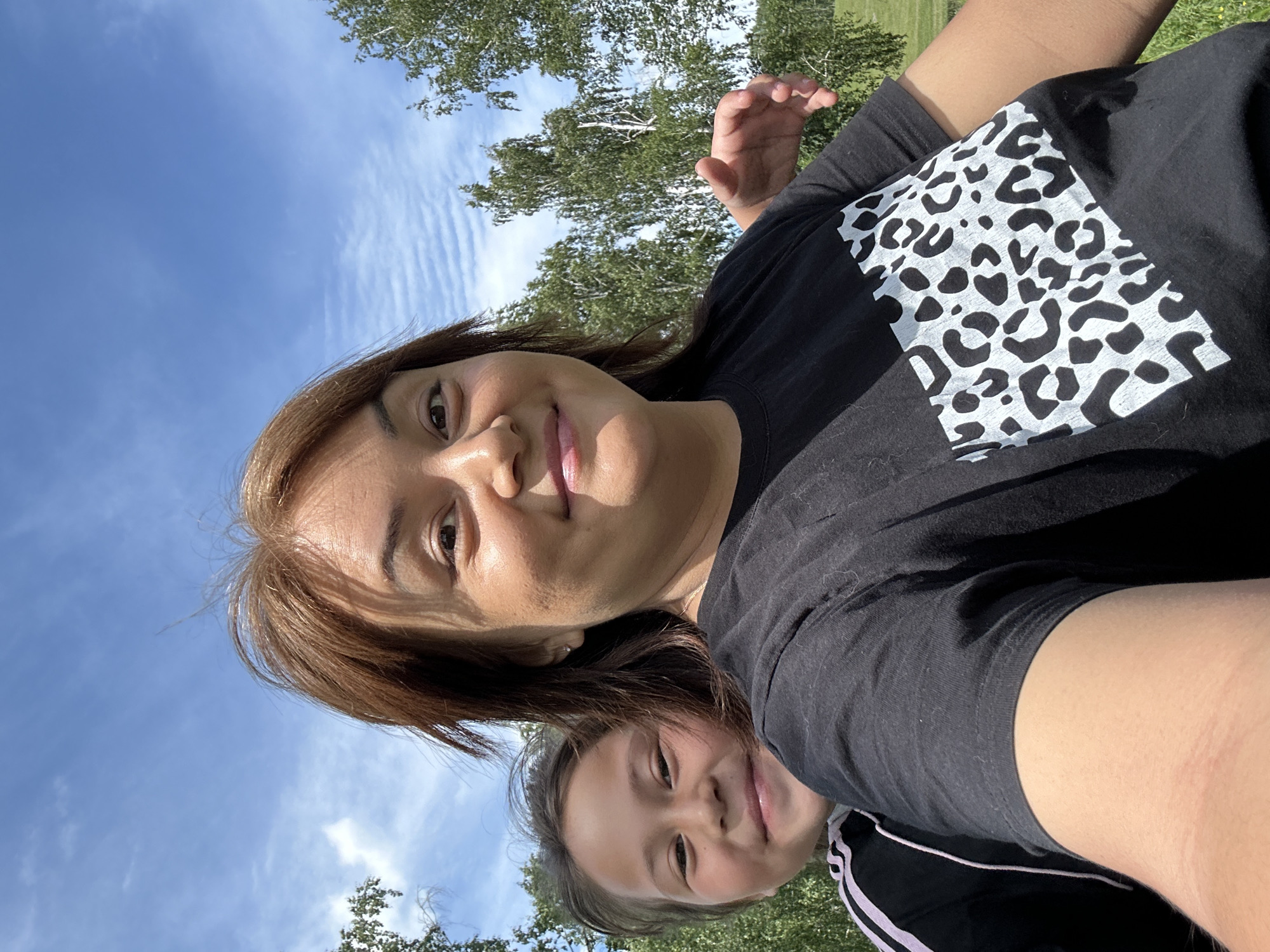 A selfie taken outdoors on a sunny day features a woman and a child from the chest up. The woman, positioned in the foreground, has brown hair and is smiling. She is wearing a black t-shirt with a white leopard print design on the chest. The child, partially visible behind the woman, is also smiling and has dark hair. The background consists of a blue sky with scattered white clouds and green trees. The location is not specified.