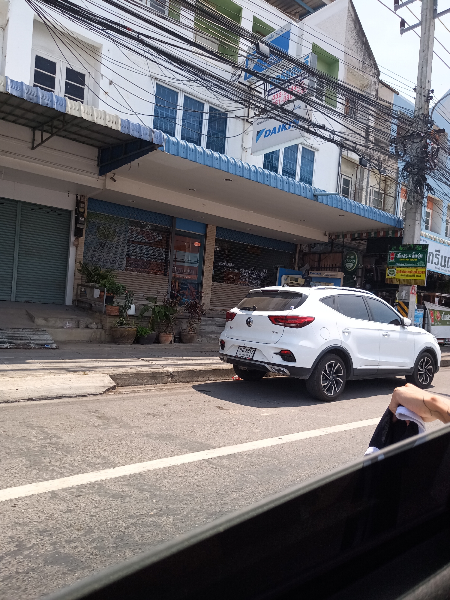 The image depicts a street scene in Hua Hin City Municipality, Thailand, as viewed from inside a vehicle. A white MG ZS car, with the license plate "กจ 5817" and "ประจวบคีรีขันธ์" visible on the plate, is parked or moving on the road.
The buildings along the street have a mix of architectural styles and are adorned with signage, including advertisements for "DAIKIN" air conditioning. There are numerous overhead electrical wires and cables crisscrossing above the street. The street appears to be asphalt and marked with white lines. There is a mix of open and closed businesses.
A person is visible within the car from which the image is taken, holding a black item with a white edge.