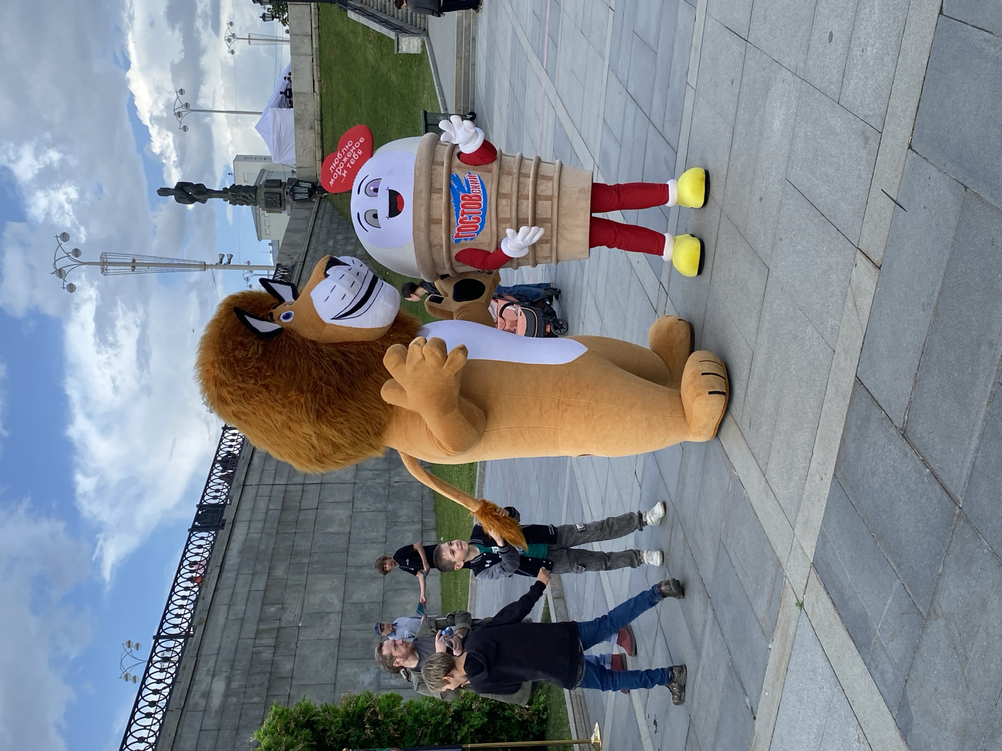 This outdoor image captures a lively scene, likely during an event or promotion in Yekaterinburg, Russia. The central figures are two large costumed mascots. On the right, a friendly ice cream cone mascot stands upright, wearing red pants, yellow shoes, and white gloves. Attached to its head is a red heart-shaped sign. On its body, the brand name "ГОСТОВский" (GOSTovsky) is visible. The red heart sign reads "люблю мороженое ...и тебя," which translates to "I love ice cream ...and you." To the left, a large, light brown lion mascot with a friendly, cartoonish face and a white muzzle is positioned, appearing to be lying down or in a relaxed pose.
Several people are scattered throughout the scene. In the foreground, a group of adults and children are observing the mascots. Two young boys are particularly engaged, looking towards the mascots; one wears a gray and black jacket, and the other a black jacket. Other adults are visible further back.
The setting is an urban public space, possibly a promenade or square, characterized by grey square paved tiles underfoot. In the background, there's a large concrete wall or structure with a black wrought-iron railing along its top. Beyond this, green grass and foliage are visible. Under a cloudy sky, several tall streetlights with multiple lamp heads are present. A monument or statue stands in the mid-ground, and a white canopy or tent is visible further in the distance, suggesting a public gathering or fair.
The weather appears to be daylight with an overcast sky, providing bright, diffused lighting. The overall atmosphere is casual and engaging, particularly for the children drawn to the mascots.
