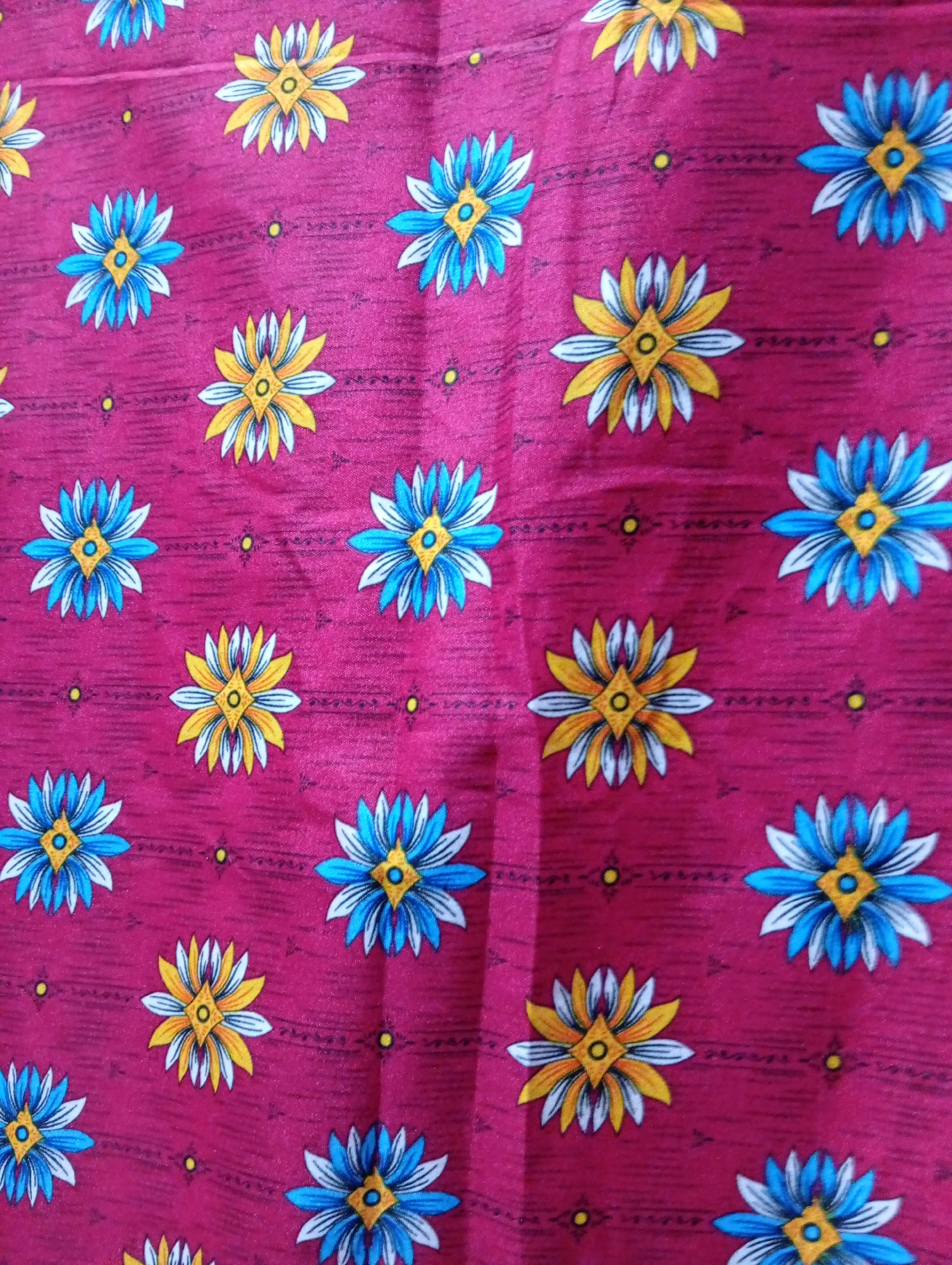 This is a close-up, top-down shot of a vibrant pink fabric with a repeating floral pattern. The flowers are stylized, with alternating blue and white petals, and a yellow center with a diamond-shaped detail. Some flowers also feature yellow and white petals with a blue and diamond center. The fabric has a subtle texture with faint horizontal lines running through it. No location in Larkana, Pakistan, is visible.