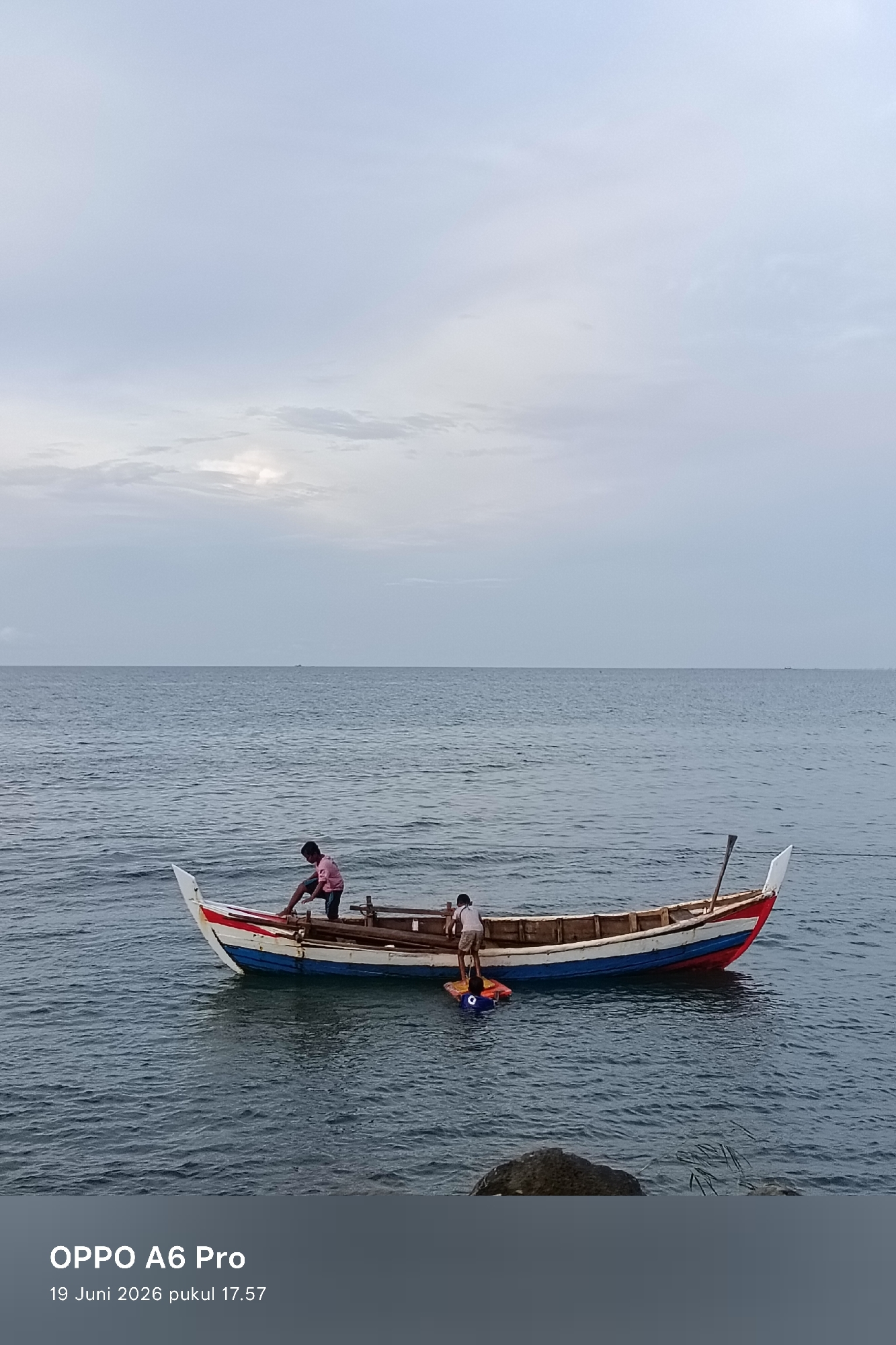 Two individuals are near a small wooden boat with a colorful hull (red, white, and blue). One person, wearing a pink shirt, is standing at the bow of the boat. Another person, in a white shirt, is in the water near the stern of the boat, interacting with a blue floatation device. The boat is floating on a body of water, likely the ocean, with a hazy sky in the background. The scene appears to be a coastal area, and given the timestamp "19 Juni 2026 pukul 17.57", it is likely from Lhokseumawe, Indonesia. The water is calm with small ripples.