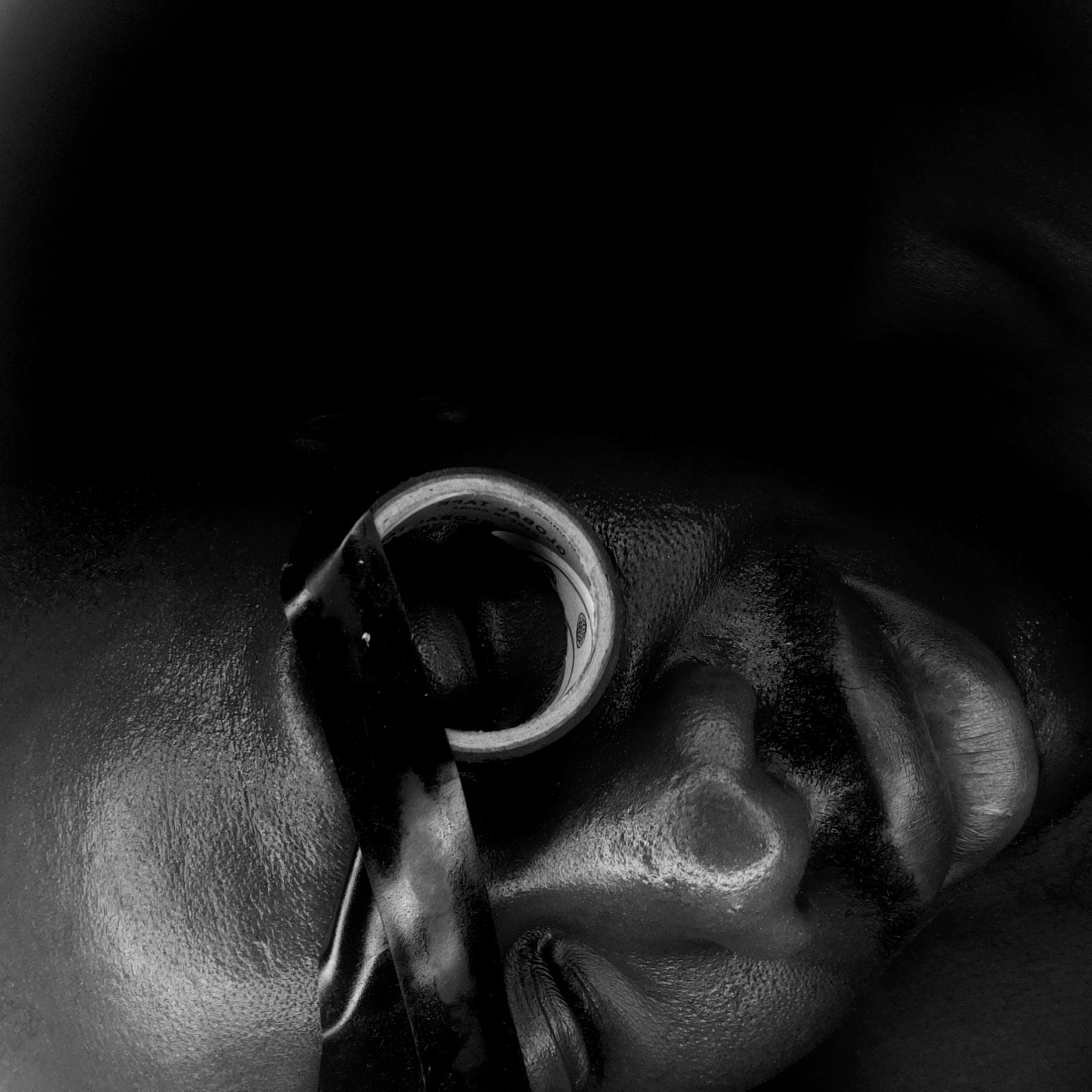 A black and white image shows a close-up of a person's face, focusing on the right ear and the area surrounding it. A roll of tape is positioned partially over the ear, with a length of black tape extending from the roll across the face. The person's skin has a glossy texture. The background is dark and indistinct. The city of Narayi, Nigeria, is not discernible from the image.