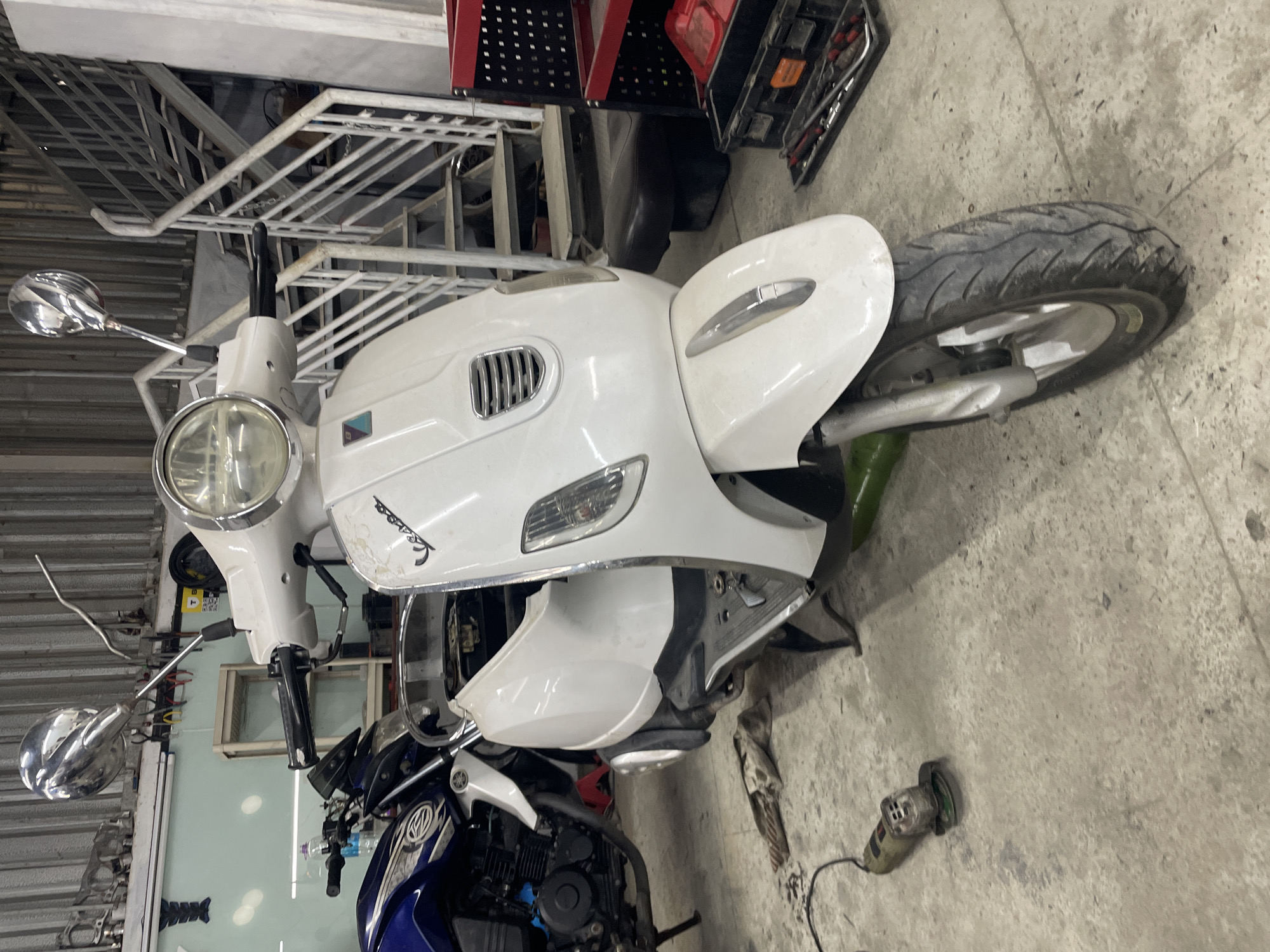 The image depicts a white Vespa scooter in Bac Nha Trang Ward, Vietnam. The scooter appears to be undergoing maintenance or repair, as parts of its body are disassembled. A blue motorcycle is positioned next to the Vespa. An angle grinder lies on the concrete floor nearby. The scene takes place within a garage or workshop environment, with metal shelving visible in the background. The overall lighting is bright, revealing details on the scooter, other equipment, and the surrounding environment.