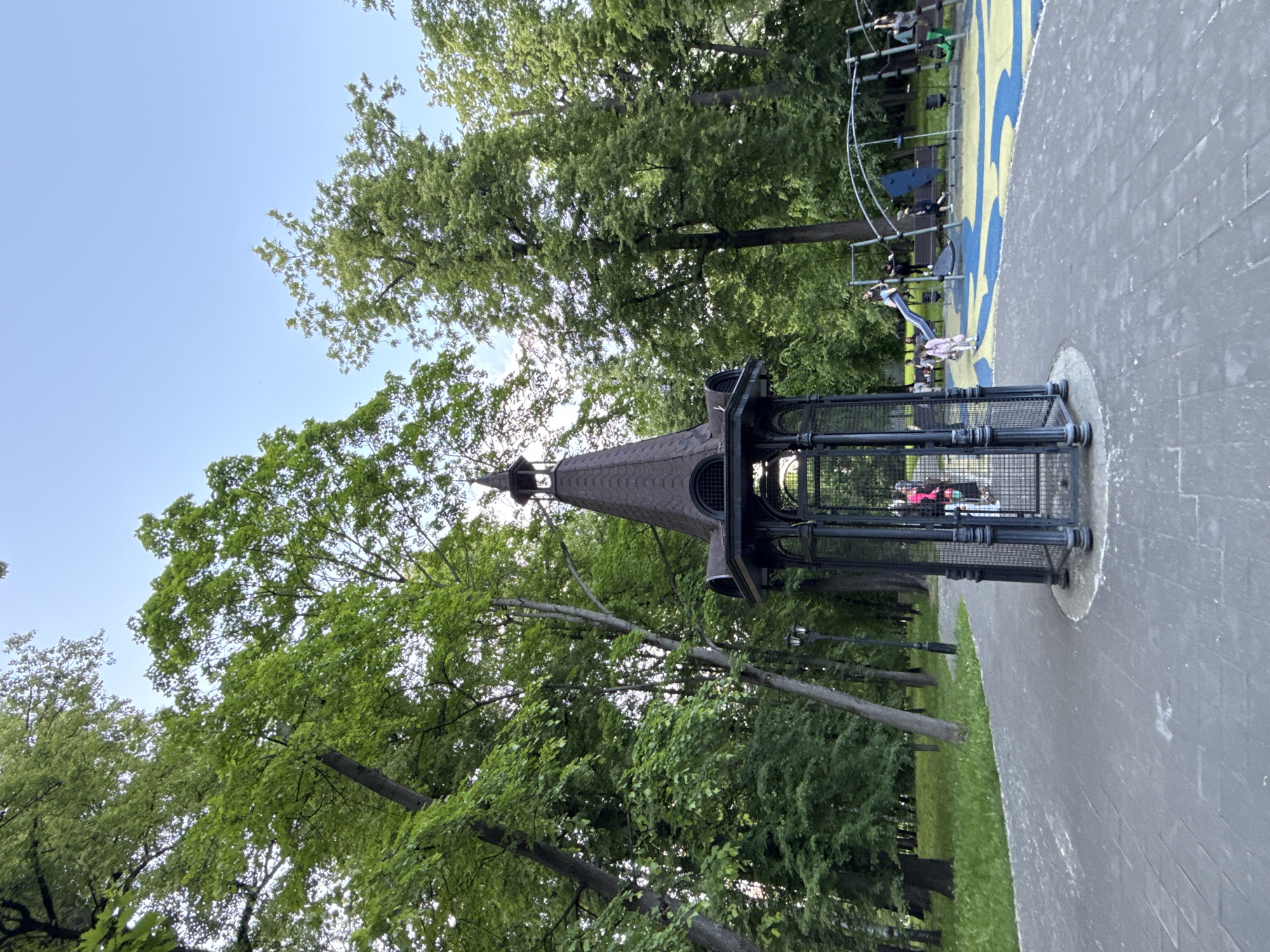 This image captures a vibrant outdoor scene in a park in Moscow, Russia, on a clear, sunny day. The foreground features a distinctive dark-colored, tower-like structure with a conical roof and grated sides, positioned on a paved surface. Adjacent to this, a modern children's playground boasts a colorful blue and yellow patterned soft floor and various climbing and sliding equipment.
Several people are engaged in activity on the playground. One person, wearing a light-colored top and dark pants, is seen descending a slide. Another person in a pink top is visible near the base of the slide, and a few other figures, likely children or supervising adults, are spread across other climbing structures. Faint figures in red and other colors can also be seen inside the tower-like structure.
The background is filled with numerous tall, lush green trees, indicating a natural and verdant setting. Above the trees, a bright, cloudless blue sky suggests pleasant weather. A paved pathway extends along the right side of the frame, separating the playground from a grassy area and additional trees. The overall scene conveys a sense of recreation and leisure during daytime. No text is visible within the image.