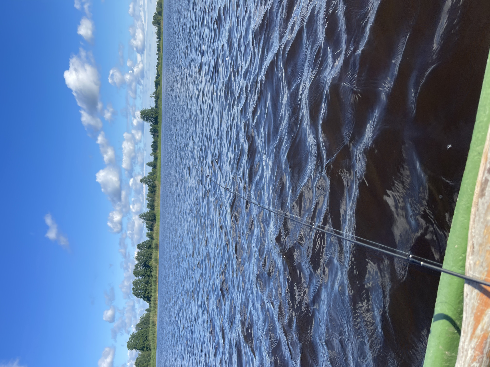 A fishing rod extends from a boat onto a dark, rippling body of water under a bright blue sky with scattered white clouds. The shoreline, lined with green trees, is visible in the distance. The foreground of the boat is partially visible and appears to be made of wood and painted green. The scene is set in Tīreļi, Latvia.
