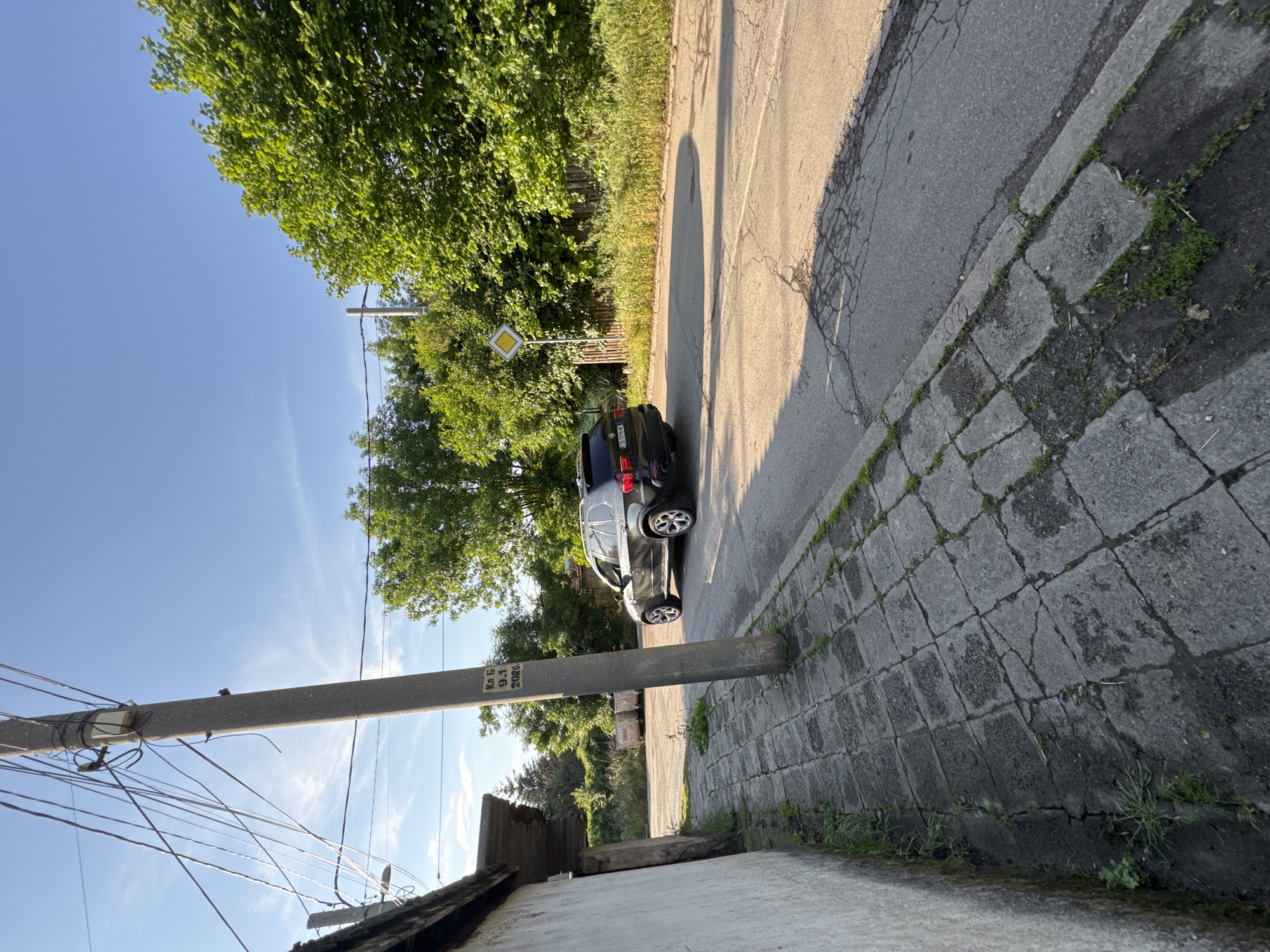 The image captures a sunlit street scene, likely in a residential or semi-rural area of Sofia, Bulgaria. The photograph is taken from a low vantage point, looking up and along a street that stretches into the distance.
In the immediate foreground on the left, a sturdy concrete utility pole rises, laden with numerous intertwined electrical and communication wires. Text visible on the pole includes "Кл Б.1", "9.1", and "2020", suggesting an identifier and a date. Behind the pole, part of a building's roof and a white wall are visible.
Adjacent to the pole, a sidewalk paved with square concrete slabs runs alongside a cracked asphalt road. Weeds and grass sprout from the cracks in both the sidewalk and the road, indicating a somewhat aged or less maintained surface.
A dark-colored SUV or crossover is parked on the right side of the road, facing away from the camera. Its rear license plate, "CB 7096 BK", is partially visible, with "CB" confirming a Sofia registration. Further down the road on the left, a yellow diamond-shaped sign, indicating a priority road, stands partially obscured by green foliage.
Lush green trees and other vegetation flank both sides of the road, casting long shadows across the asphalt, suggestive of either early morning or late afternoon sunlight. One prominent oval-shaped shadow extends across the road from an unseen object. The sky above is a clear, vibrant blue with a few subtle wispy clouds. The overall impression is one of a bright, peaceful, and sunny day. No people or discernible activities are present in the scene.