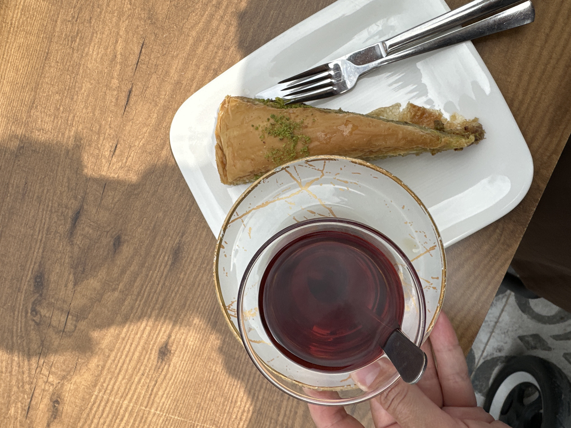 This high-angle, close-up image captures a delightful moment of refreshment on a wooden table, likely in Fethiye, Turkey. In the foreground, a person's hand gently holds a clear glass teacup, filled with dark red Turkish tea, which rests on an elegant white saucer adorned with gold, marbling-like patterns.
Adjacent to the tea, a single triangular piece of baklava, liberally sprinkled with green pistachio crumbs, is presented on a minimalist white rectangular plate. A set of polished silver cutlery, a fork and a knife, are neatly arranged on the plate, positioned above the pastry.
The wooden table surface, characterized by its warm brown tones and visible grain, is illuminated by strong natural light, casting distinct shadows across its surface and suggesting it is daytime. A small portion of a patterned, possibly tiled, floor is visible in the bottom right corner of the frame. The scene evokes a sense of relaxation and enjoyment, typical of a casual cafe setting or a pleasant break. There is no visible text in the image.