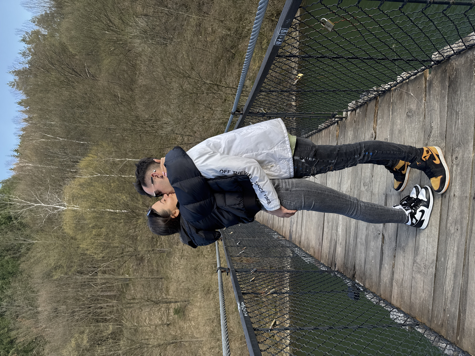 A couple is kissing on a wooden suspension bridge in Dolni Pasarel, Bulgaria. The woman has dark hair and is wearing a black puffer jacket and grey jeans. The man has dark hair, glasses, a white jacket with "OFF THE WALL SINCE 1966" written on the sleeve, black jeans, and tan/black sneakers. They are standing on the bridge, which has a metal safety fence with locks attached to it. A lake or body of water is visible beneath the bridge, along with a background of leafless trees.