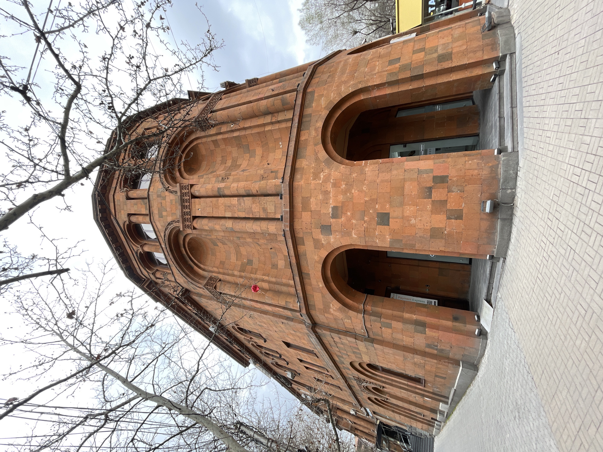 The image captures the architectural detail of the Yerevan City Hall, located in Yerevan, Armenia. The building is constructed from reddish-brown stone with a distinct arched facade. The angle emphasizes the building's height and form, with bare tree branches in the foreground. The city hall is situated on a paved walkway with a cloudy sky in the background.