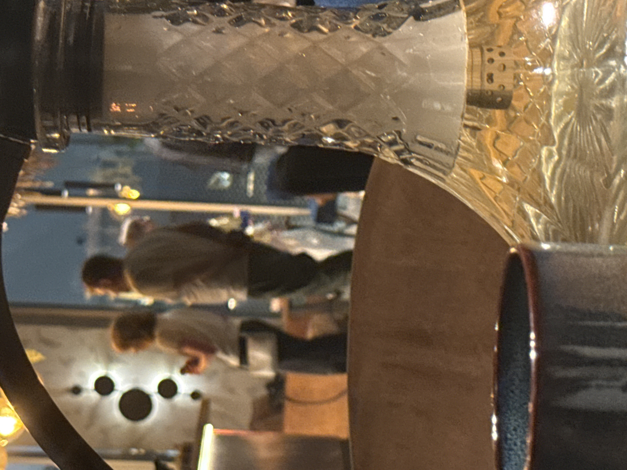 The image captures an indoor scene, likely a bar or lounge, with a blurry background suggesting an urban environment at dusk or night. In the foreground, a portion of a hookah is visible, its glass base filled with liquid and featuring a diamond-patterned texture. To the left, the rim of a dark-colored cup is also in focus. The middle ground shows two individuals standing and facing away from the camera. The person on the left, with reddish hair, appears to be holding a mobile phone. The person on the right is a man in a light-colored shirt. In the far background, a blurred cityscape with lights is visible, possibly indicating a location within the city of Bratislava, Slovakia. The overall ambiance is dimly lit, with artificial lights contributing to the scene's illumination.