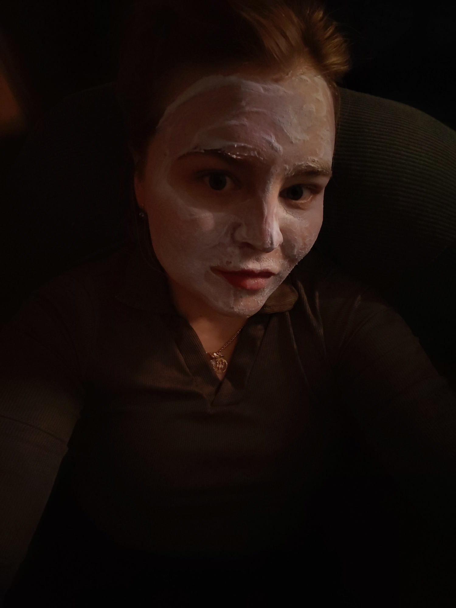 A woman is seen from a close-up, low-angle perspective. She has a white facial mask applied to her face, covering her forehead, nose, cheeks, and chin. Her hair is light brown. She wears a dark-colored, V-neck top. A gold necklace is visible. The background is dark and indistinct. There are no indications in the image that would allow one to determine that the location is in Izhevsk, Russia.