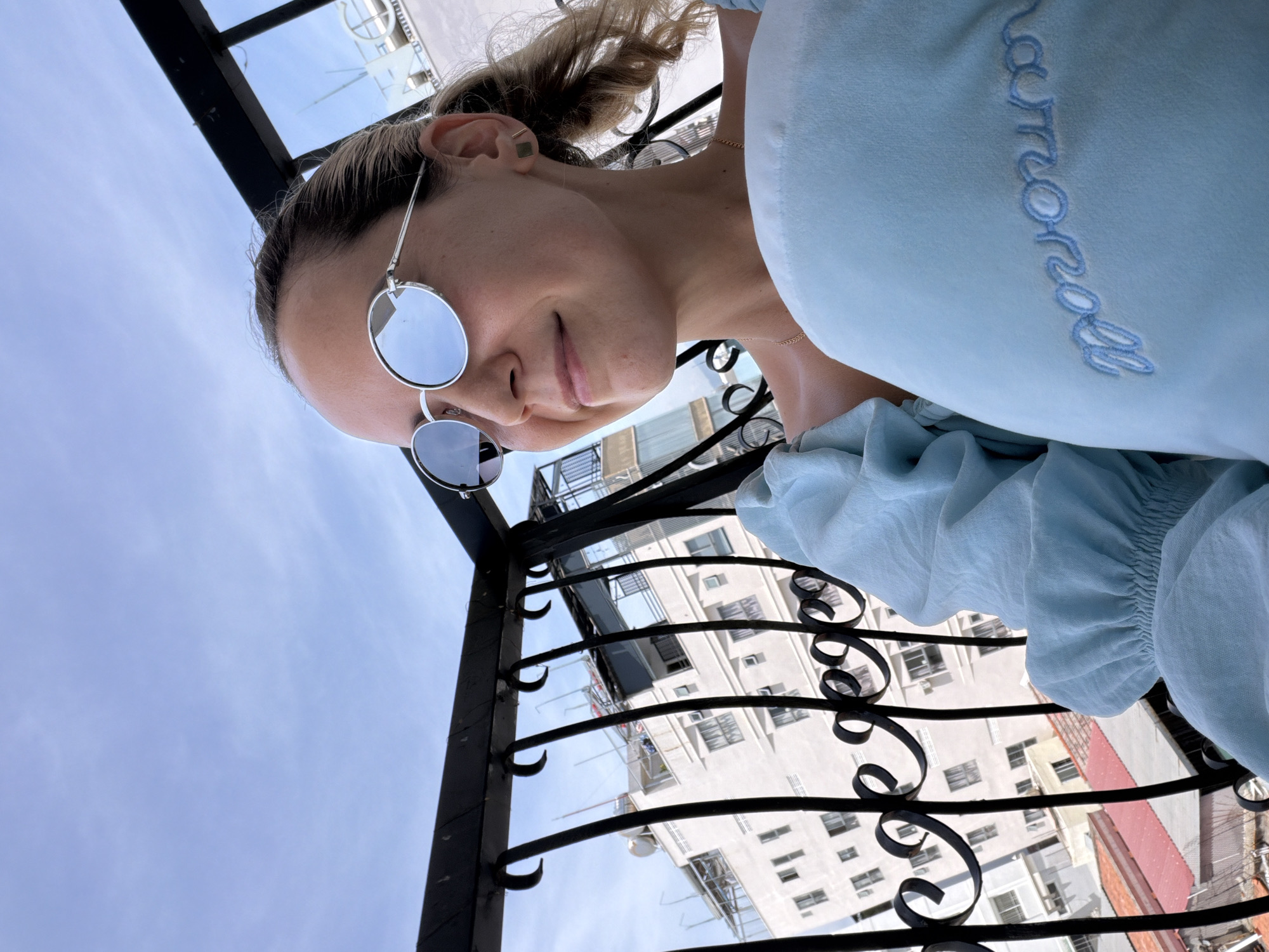 A young woman with blonde hair pulled back in a ponytail stands on a balcony overlooking a city. She is wearing round, silver-rimmed sunglasses and a light blue top. She holds a light blue plush toy in her arms, which has embroidered text on it. The balcony railing is black and ornate. In the background, several buildings of varying heights are visible, with some showing signs of construction or renovation. The sky is mostly clear with a few wispy clouds. The scene is consistent with an urban environment in Nha Trang Ward, Vietnam.