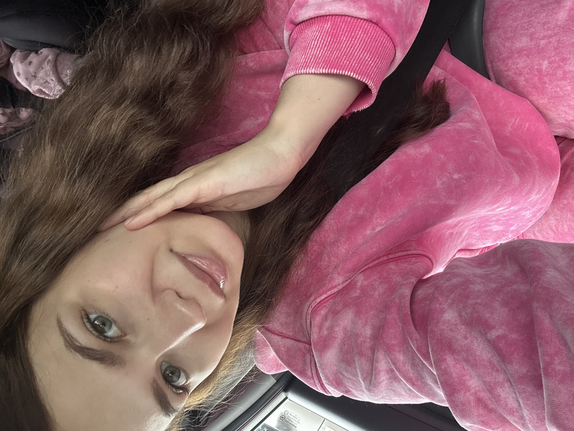 A woman with long brown hair and green eyes is taking a selfie in a car. She is wearing a pink tie-dye sweatshirt. Her left hand is resting on her face, and her right arm is bent. The image is taken indoors, likely during the daytime. The woman appears relaxed and happy. There is some visible text on a sticker or label in the lower right corner of the image, but it is too blurry to read.