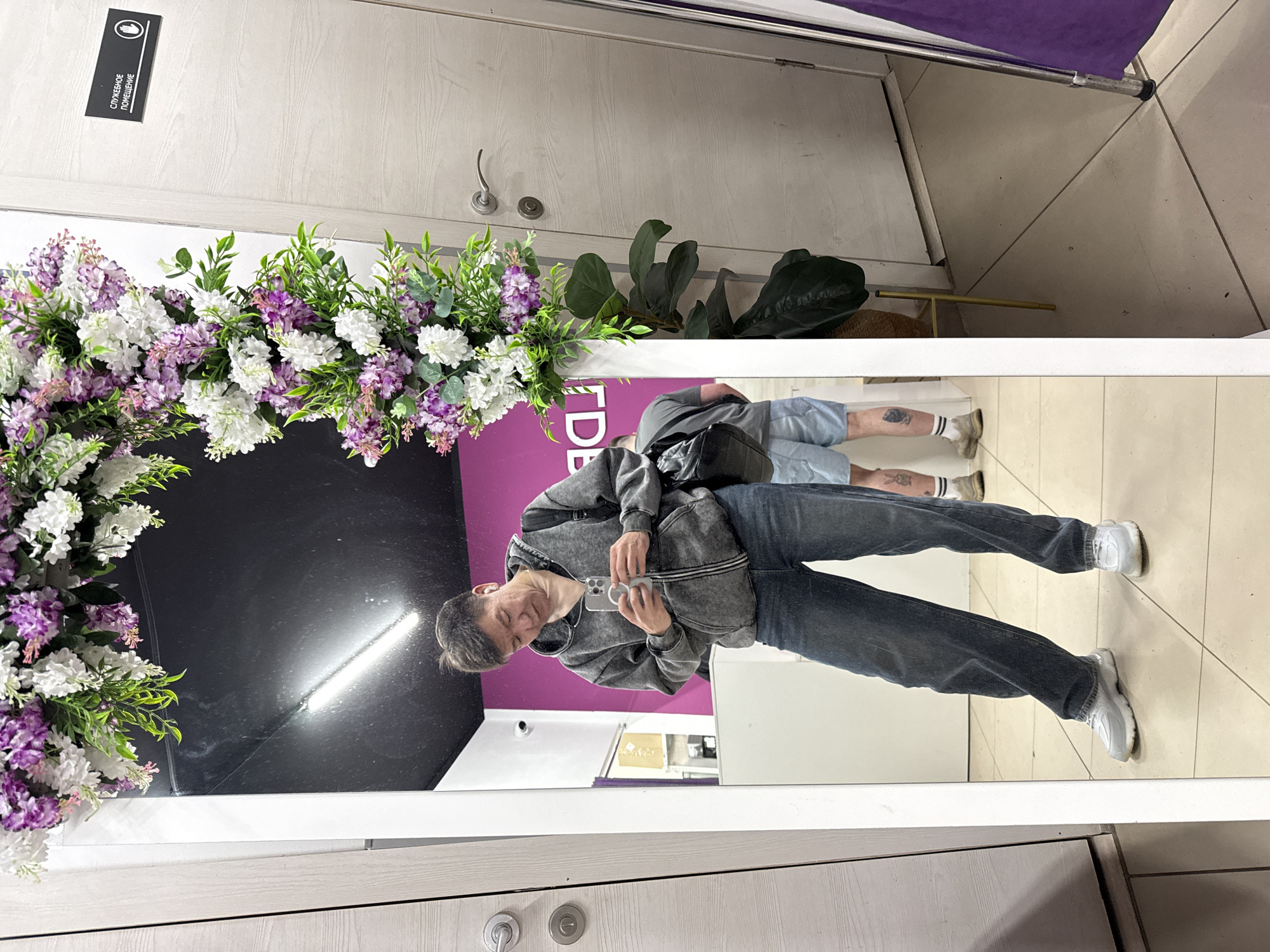 The image captures a mirror selfie of a person in Moscow, Russia. The reflection shows a man holding a smartphone, wearing a grey hooded jacket and jeans. Behind him in the mirror, another person in shorts is also visible. The mirror is decorated with artificial white and purple flowers. A sign in Russian is mounted on the wall. In the background, clothing racks and flooring tiles are visible, indicating the setting is likely a clothing store.