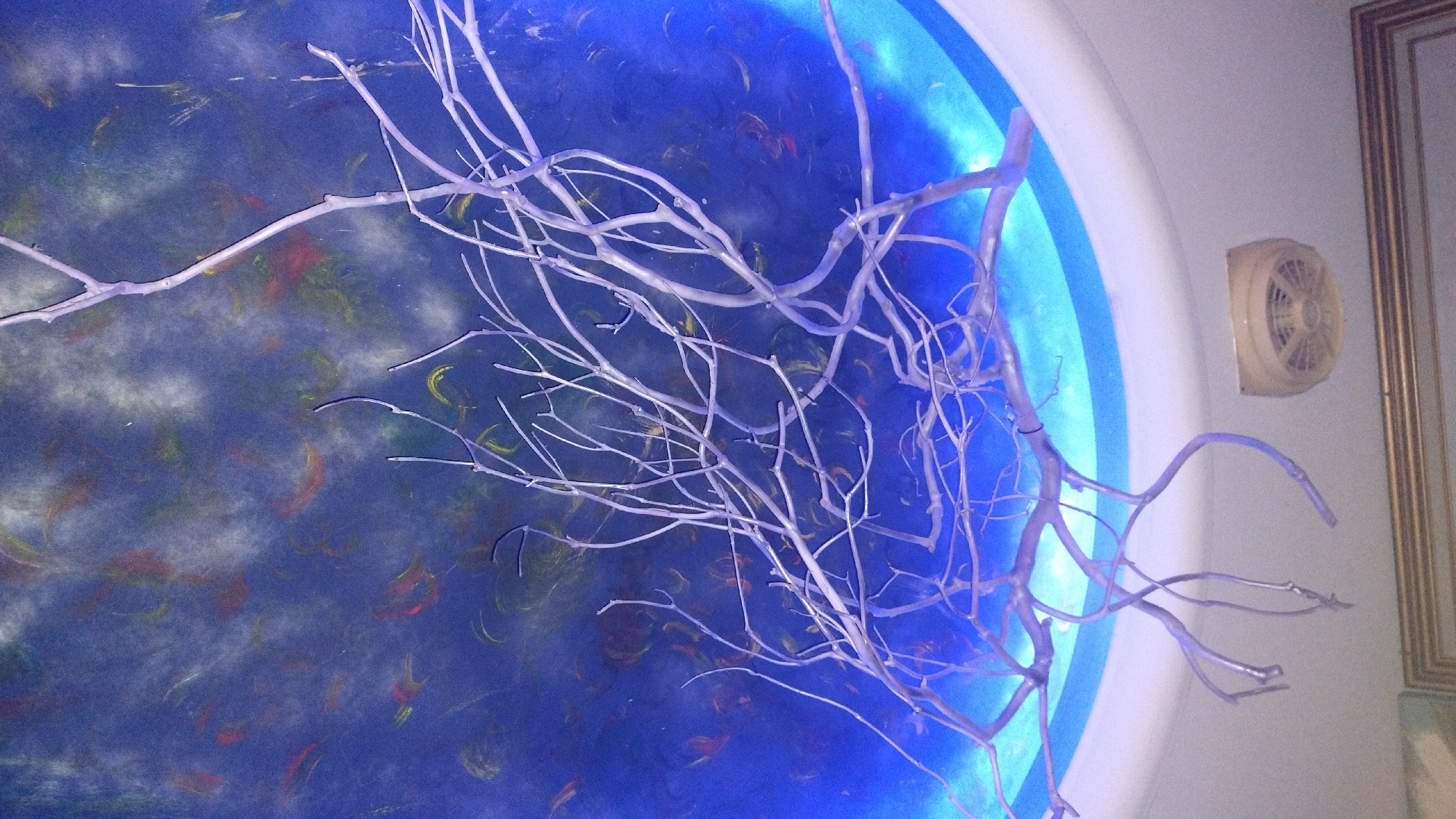 This image shows a close-up of a white, bare branch art installation. The branch is detailed with many smaller twigs and appears to be made of a painted material. It is positioned in front of a background that seems to be a wall or ceiling decorated with abstract shapes in shades of blue, red, and yellow. The lighting suggests an indoor environment, possibly with ambient blue lighting emanating from behind the painted surface. To the right of the branch, there is a beige ventilation grate mounted on a light-colored wall. Further to the right, a framed picture with a gold frame is visible, leaning against the wall. The overall scene evokes a sense of artistic display or interior decoration. There is no visible text in the image.
