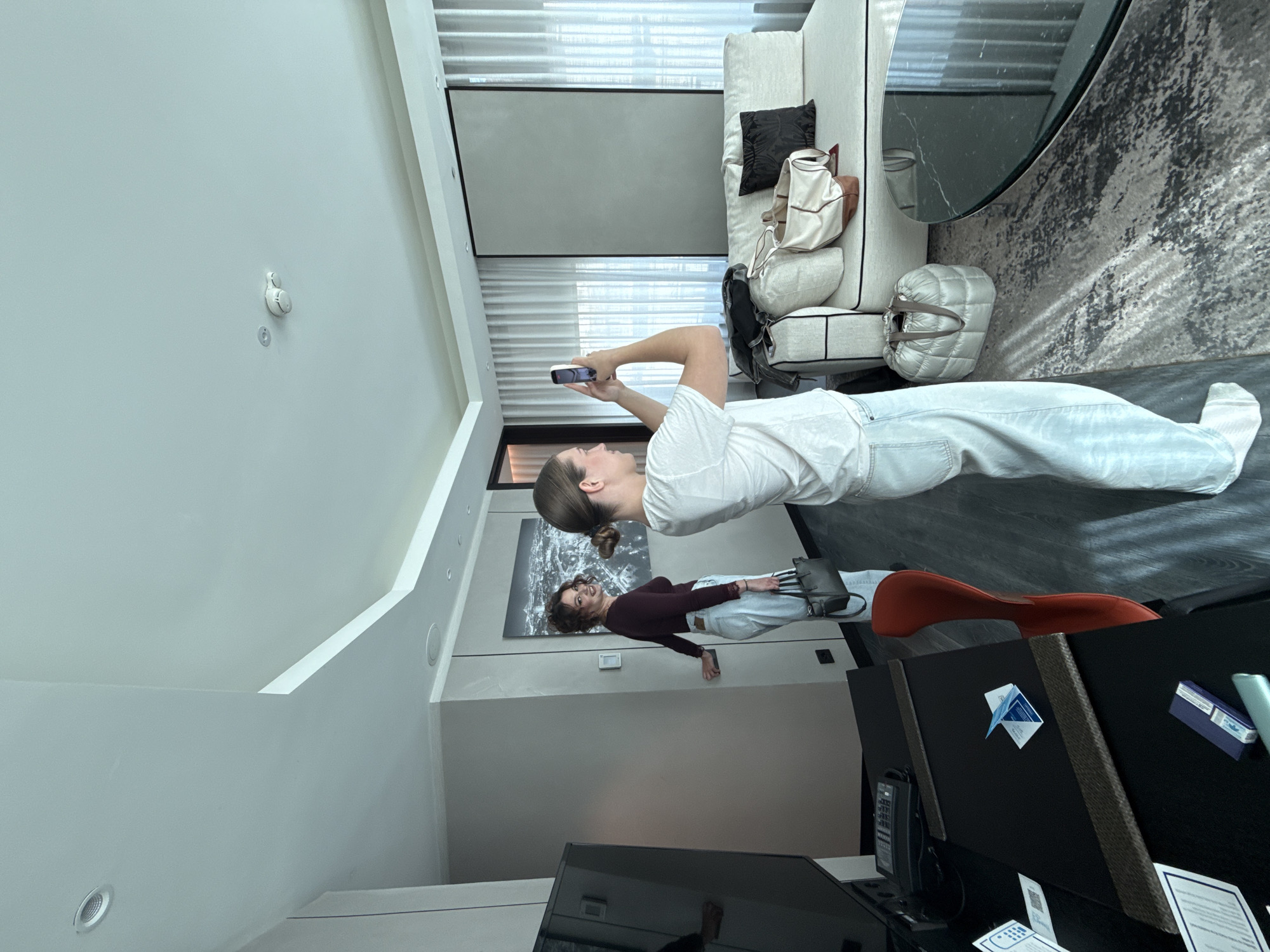 The image depicts a modern living room interior. Two women are visible. One is standing and taking a photo with her smartphone of the other woman, who is lying down. The room contains a sofa with pillows and bags, a round table, a rug, a chair, and a TV. The wall art is also visible. The setting resembles a residential or hotel interior in Vracar Urban Municipality, Serbia.
