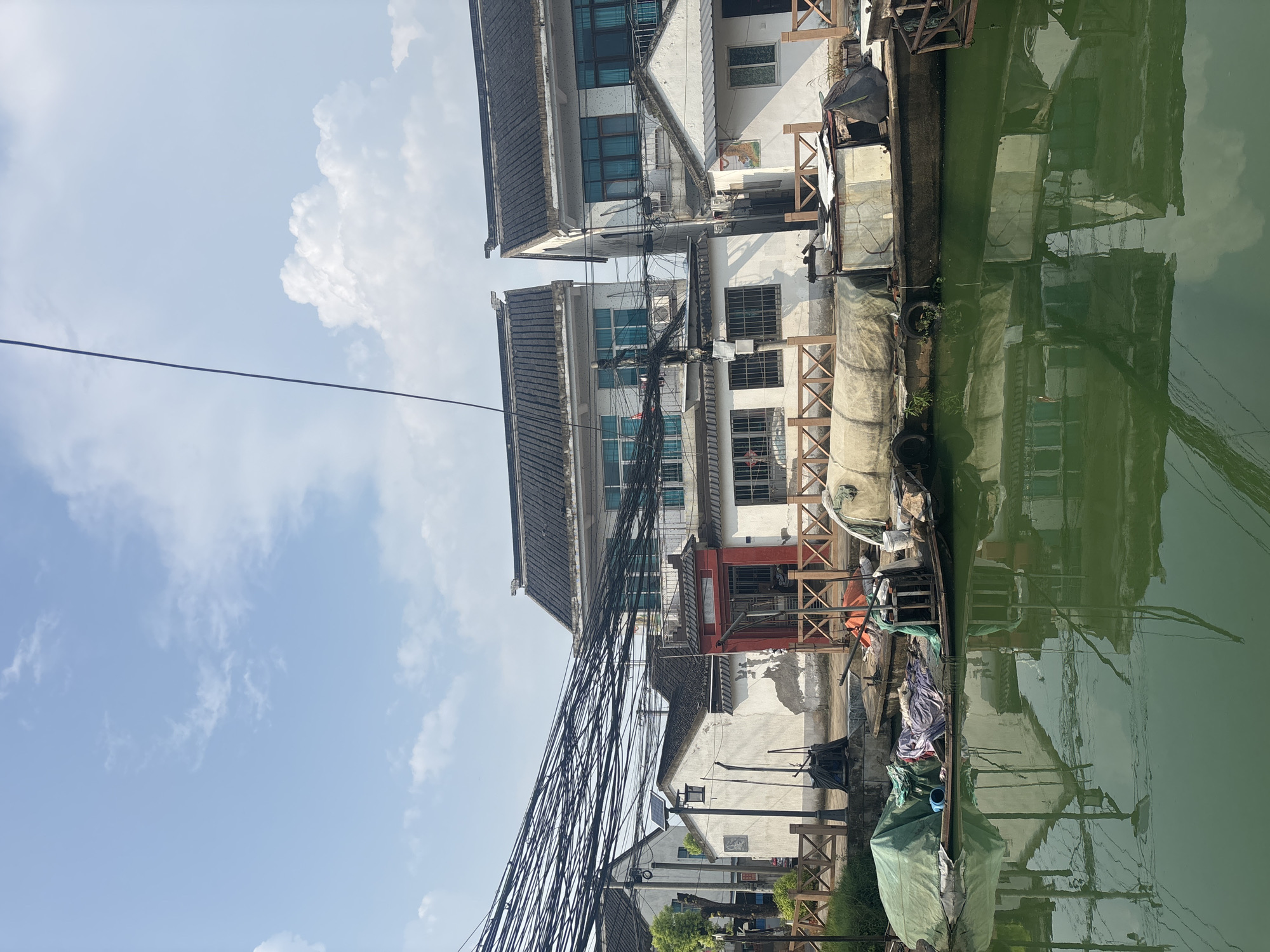 The image depicts a canal scene in Wuzhong District, China, under a partly cloudy sky. Traditional white buildings with dark tiled roofs line the canal. A dense network of black electrical wires stretches across the upper portion of the image, obscuring a significant portion of the sky and buildings.
In the foreground, a narrow, dark green canal occupies the lower half of the frame. The water's surface is somewhat murky, reflecting the buildings and the sky. A long, narrow boat is moored along the bank. The boat is laden with various objects covered by tarps, including what appears to be a green tarp on the left, several fabric bundles in the center, and other materials. One section of the boat's deck is uncovered, revealing weathered wood and some metal structures. Tires are visible along the side of the boat.
Along the canal bank, a wooden fence with crisscrossing slats separates the water from the buildings. Behind the fence, the white walls of the buildings show some signs of wear and tear. One building features a prominent red doorway with intricate detailing and grilled windows. Other buildings have multiple windows, some with blue-tinted glass. A street lamp with a solar panel is visible near one of the buildings.
The architecture suggests a traditional Chinese style, common in areas with canals. The overall impression is one of a working canal with moored boats and functional, albeit somewhat aged, buildings. No people are explicitly visible in the scene, but the presence of the boat and stored items implies human activity or habitation. The electrical wires suggest a modern utility infrastructure overlaid on a more traditional environment.